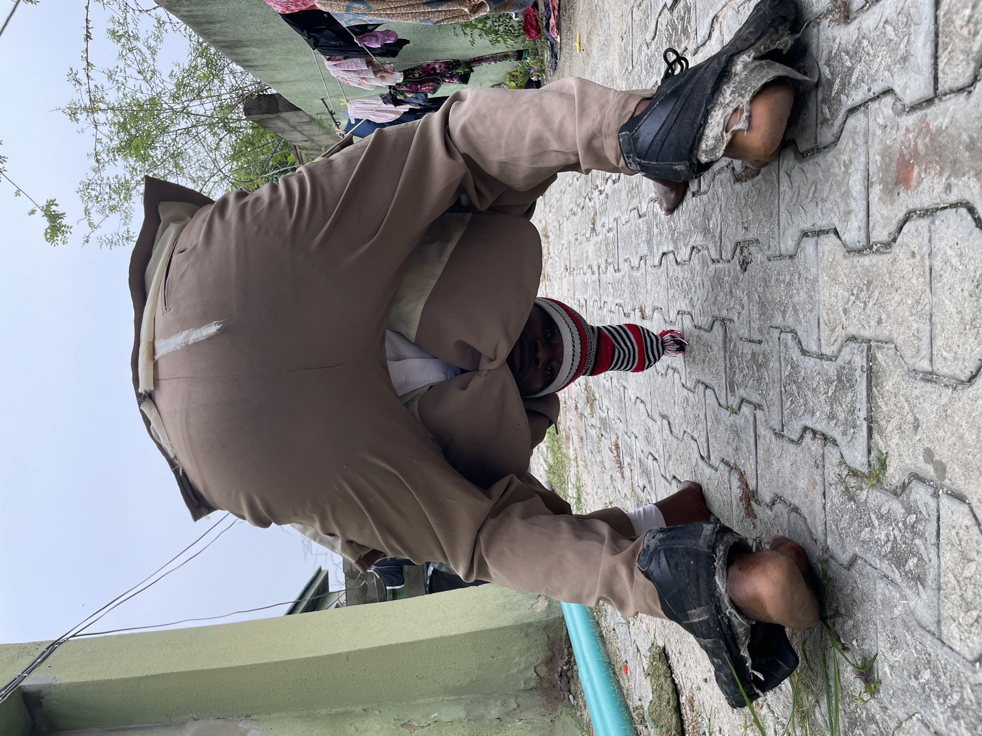 A person wearing khaki trousers, a white shirt, and a red and white striped beanie is crouching on a concrete-paved area. They are wearing severely damaged black shoes, with their bare heels exposed. The person is looking directly at the camera. Overhead, clothes are drying on a line, and green foliage is visible. The location is believed to be Ado, Nigeria. A light green concrete structure runs along the lower left of the frame. A blue pipe is visible next to the green concrete structure. The concrete pavement is made of interlocking hexagonal blocks.