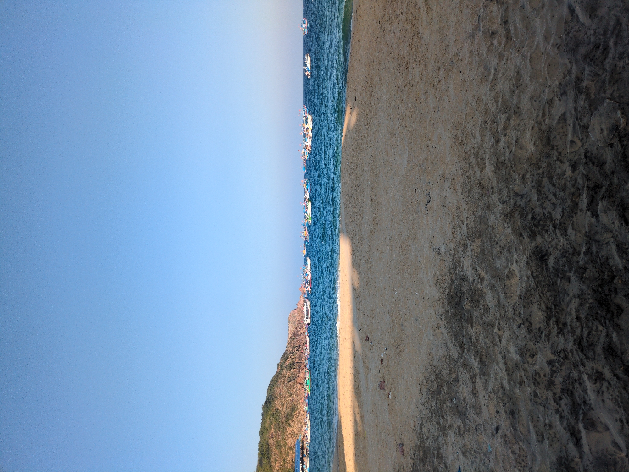 This image captures a coastal landscape in East Quy Nhon Ward, Vietnam, under a clear sky. The upper third of the frame shows a vast, unblemished light blue sky. The middle section is dominated by a body of blue-green ocean with small, visible waves. Approximately 20 to 25 boats are moored or anchored across the water, extending horizontally from left to right. These boats are varied in size and color, predominantly white with blue, green, and red accents.
On the left side of the scene, a prominent, rugged hill or small mountain covered in green vegetation meets the ocean. A small structure with a blue roof is discernible at the base of this landmass on the far left. The foreground consists of a sandy beach, displaying varying tones of light tan to dark brown, suggesting differences in moisture content or sediment composition. Scattered minor debris is visible on the sand. A distinct, elongated shadow stretches diagonally from the lower left across a significant portion of the beach. No individuals are present, and no specific actions or interactions are depicted within the frame.