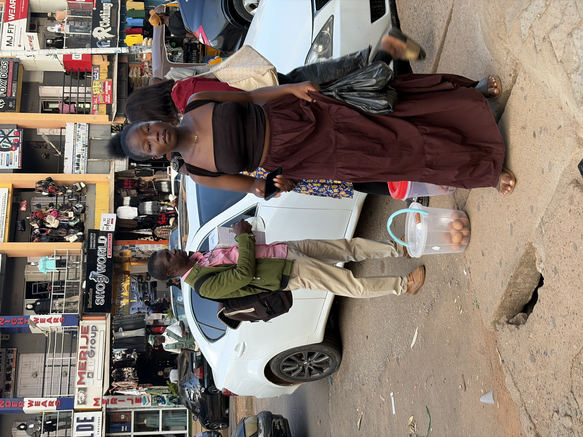 This image captures a street scene in Jos, Nigeria, featuring several individuals and commercial establishments. In the foreground, a young woman wearing a dark brown strapless top and a long, flowing maroon skirt stands facing the camera, holding a black mobile phone in her right hand and a black plastic bag in her left. To her left, a man in a green jacket over a pink shirt and tan trousers is seen from the side, holding a box and looking towards his right. Beside him, to the right, is a white sedan. A clear plastic bucket containing several eggs is positioned on the ground near the man's feet. In the background, a multi-story building houses various shops. Signs visible include "MERIJE Group", "SHOE WORLD WHOLESALES", and "MJ FIT WEAR". People are seen browsing in front of the shops. The environment appears to be an urban street with vehicles and pedestrian activity.