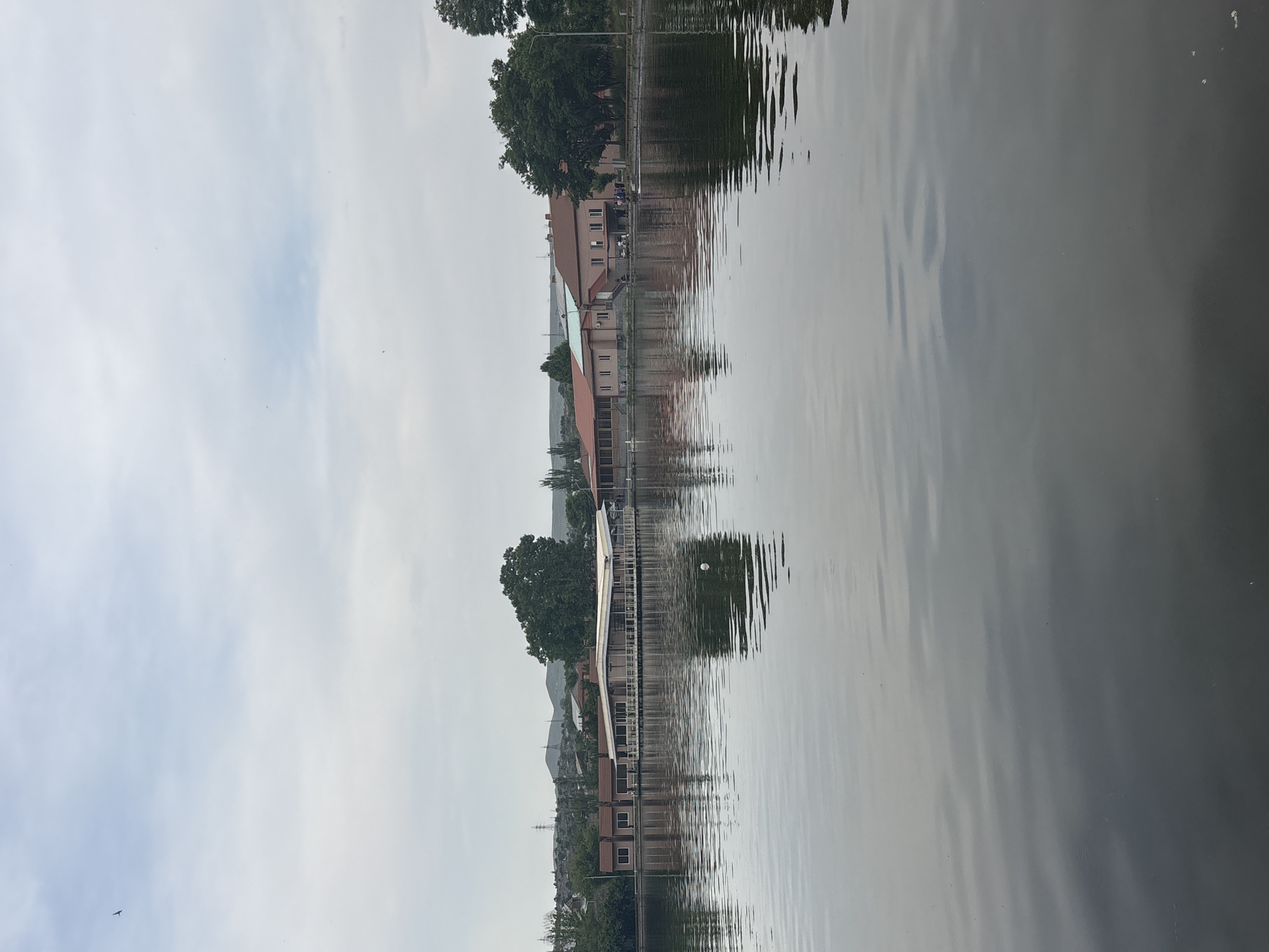 The image depicts a body of water, likely a river or lake, with reflections of buildings and trees. The scene is in a somewhat overcast condition, and the water surface is rippled, blurring the reflections. On the far shore, there are buildings with various architectural styles. Some of the buildings appear to be residential, while others may be commercial or public structures. There are trees interspersed among the buildings, contributing to the landscape. The sky is visible in the upper part of the image. I can say this is the city of Yerevan, Armenia, because the architecture of the buildings in the image seems to be consistent with Yerevan.
