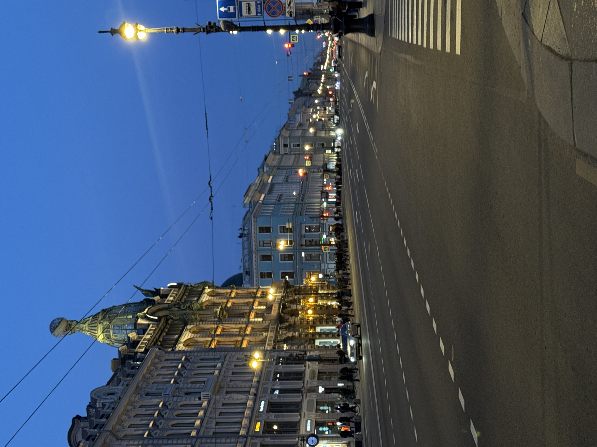 The image depicts a street in Saint Petersburg, Russia at dusk. A wide boulevard runs along the right side of the frame, marked with white dashed lines and a pedestrian crosswalk. On the left, ornate buildings line the street, illuminated by streetlights and building lights. The Singer House, topped with a glass dome, is visible among the buildings. Streetlights extend above the buildings, with wires stretching across the sky. People can be seen walking on the sidewalk in front of the buildings. A few vehicles travel down the road. Traffic signs on poles are also visible on the upper left. The sky is a gradient of dark blue.