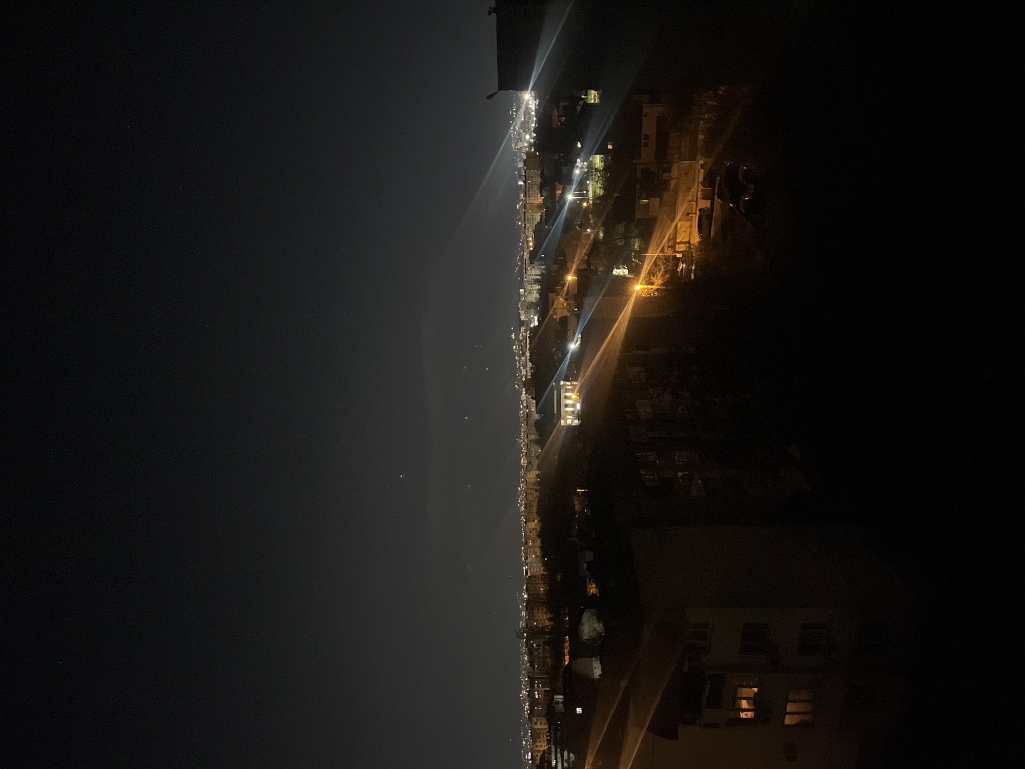 The image depicts a nighttime view of Sofia, Bulgaria, from an elevated vantage point. A clear sky is visible on the left side of the image, transitioning into a cityscape with numerous lights. Buildings and streetlights are scattered throughout the city. Some light beams are present, suggesting headlights from vehicles traveling along the streets. Buildings are visible in the foreground on the right side of the image. The scene appears dark overall with the city lights illuminating certain areas.