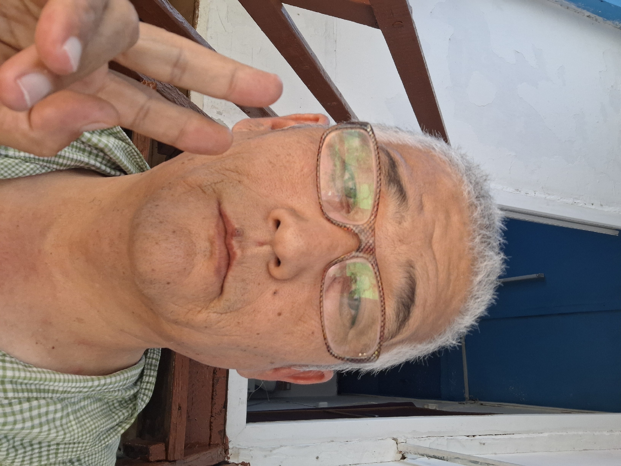 In a close-up, eye-level selfie, an East Asian man with grey hair, glasses, and a pale complexion poses with his right hand held up, fingers forming a peace sign. He is wearing a green and white checkered shirt. The background suggests an outdoor setting, possibly near a building with weathered white walls and dark wooden railings and steps. The sky above is a bright blue, partially obscured by the roofline of an overhanging structure. The man's face is the primary focus, with his eyes visible through his glasses and a neutral expression. The overall impression is casual and candid. The image does not provide any specific visual cues to determine its location within Almaty, Kazakhstan.