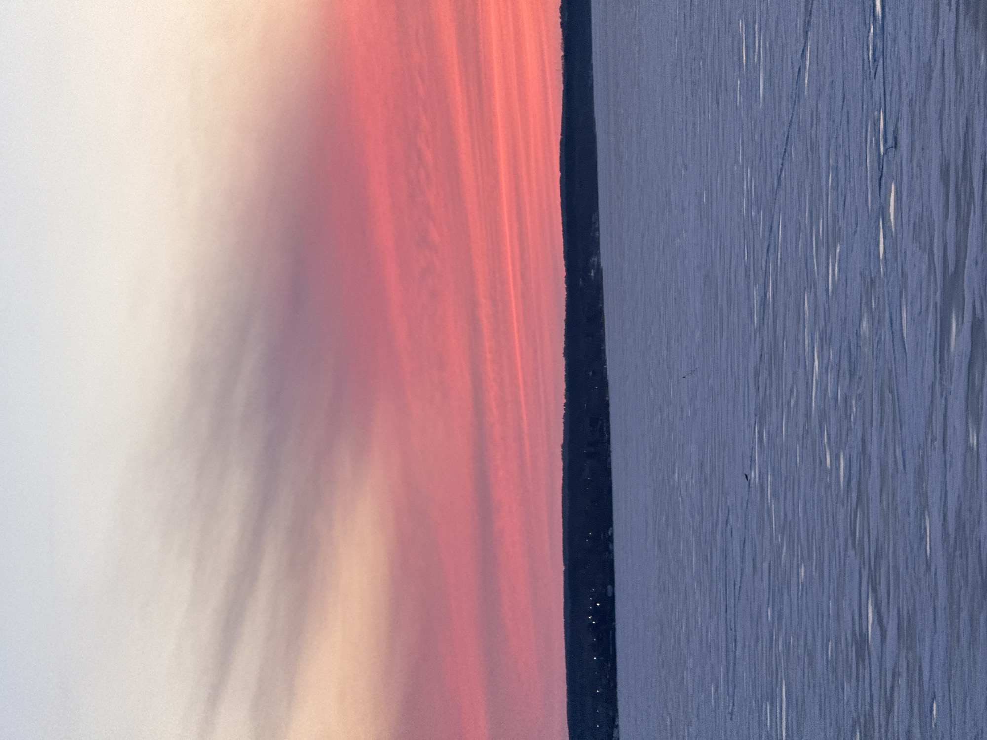 The image shows a horizontal view of what appears to be a body of water, likely the Votkinsk Reservoir, with a dark shoreline separating it from a sky with a striking sunset. The sky displays gradations of pink, orange, and white hues. The water exhibits a textured surface, suggesting either a ripple effect or ice formation. Small, sparse light sources are visible along the shoreline, likely indicating buildings or structures within the city of Votkinsk. The overall composition is simple and serene.