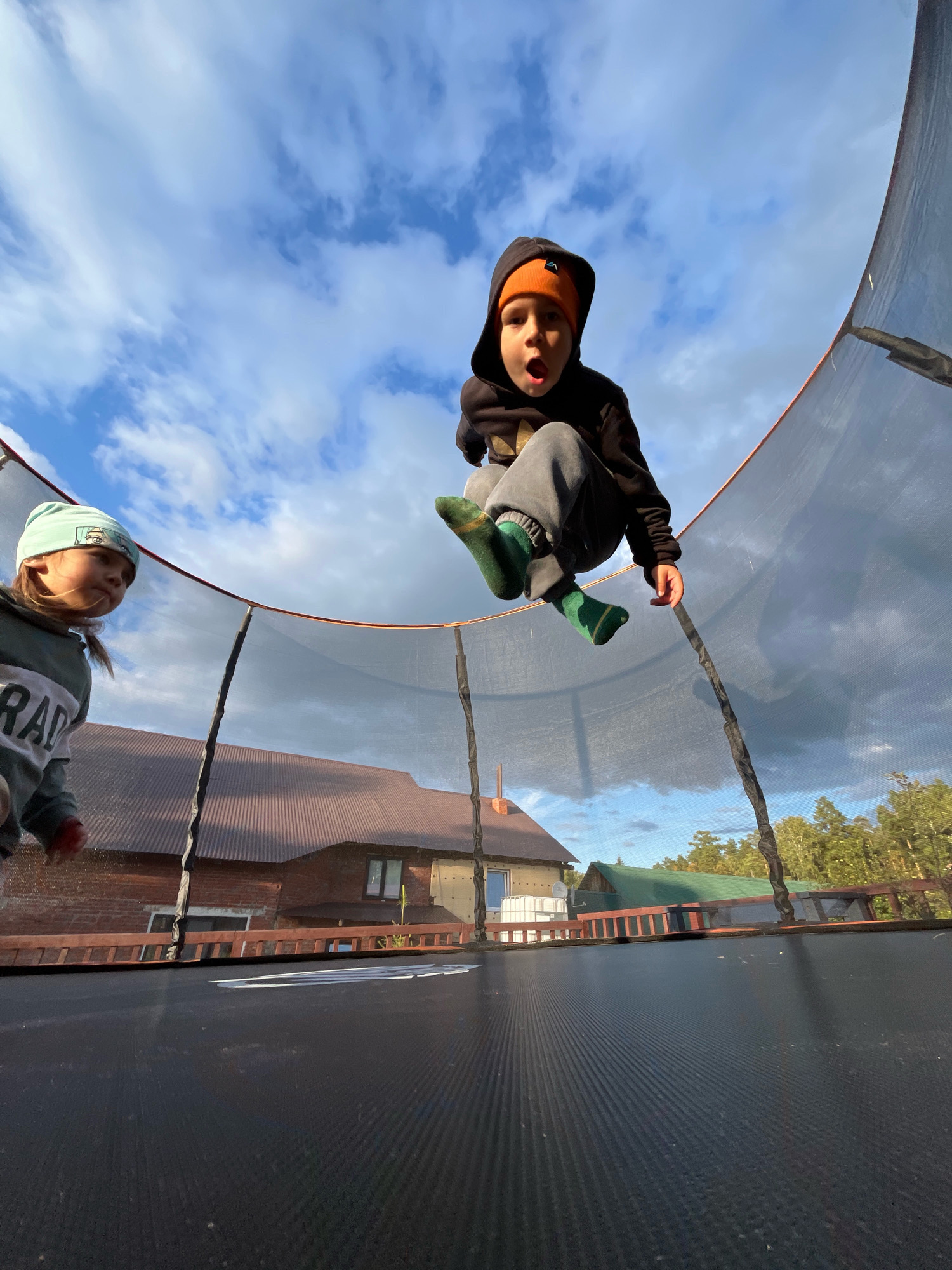 In Chaikovsky, Russia, two children are jumping on a trampoline. A boy in a black hoodie and orange beanie is mid-air, his mouth open in surprise and his tongue sticking out. He is wearing grey sweatpants and green socks. To his left, a girl in a light blue beanie and grey sweatshirt is looking at him. The trampoline has a black mat and a black safety net. In the background, a brick house with a brown roof is visible, along with a green barn and trees. The sky is blue with white clouds.