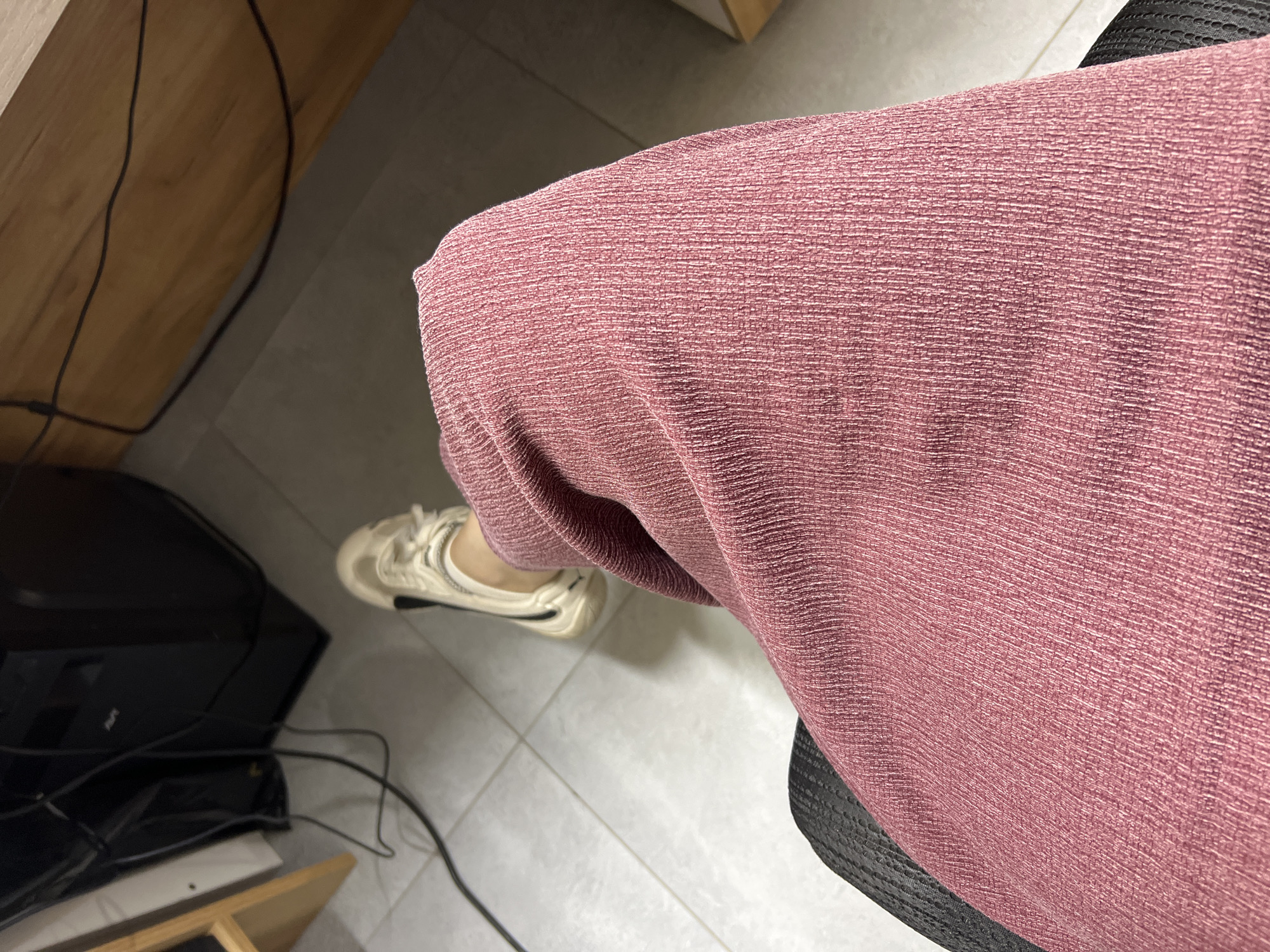 The image shows a close-up view of a person's legs, covered by a pink, textured garment, and a white sneaker. The person is seated in a black chair. In the background, a black computer tower and various cables are visible on a light-colored tiled floor. A wooden surface is also present in the upper right portion of the image. The scene appears to be indoors, possibly in an office or study. There is no information in the image to suggest a location in Shahrixon, Uzbekistan.