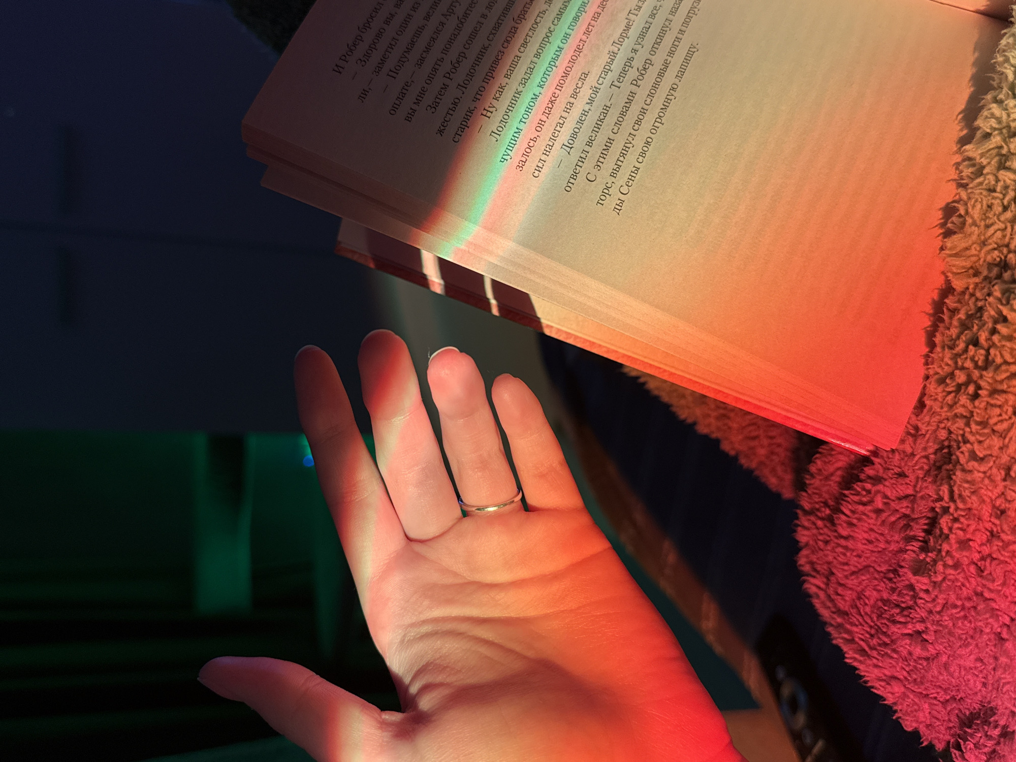 A close-up, high-angle indoor shot features a human hand with a silver ring on the fourth finger, prominently displayed in the foreground. The palm of the hand faces upwards, illuminated by a striking gradient of red, orange, and yellow light. This vibrant, multi-colored light, reminiscent of a rainbow or prism effect, also falls across an open book positioned in the upper right and center. Bands of green, blue, and yellow light are visible across the left page of the book, transitioning into warmer reds and oranges on the right page.
The book contains visible Russian text, including phrases such as "И Робер бросил" (And Robert threw), "Лодочник задал вопрос самым" (The boatman asked a question in the most), and "ответил великан" (the giant replied). To the right of the book, a textured, fluffy object, possibly a blanket or piece of furniture, is visible, saturated in a reddish-brown hue by the warm light. The background on the left is dark and blurred, with subtle hints of green and blue. The overall scene suggests a moment of quiet activity, possibly reading, in a cozy indoor setting, bathed in atmospheric and colored lighting.