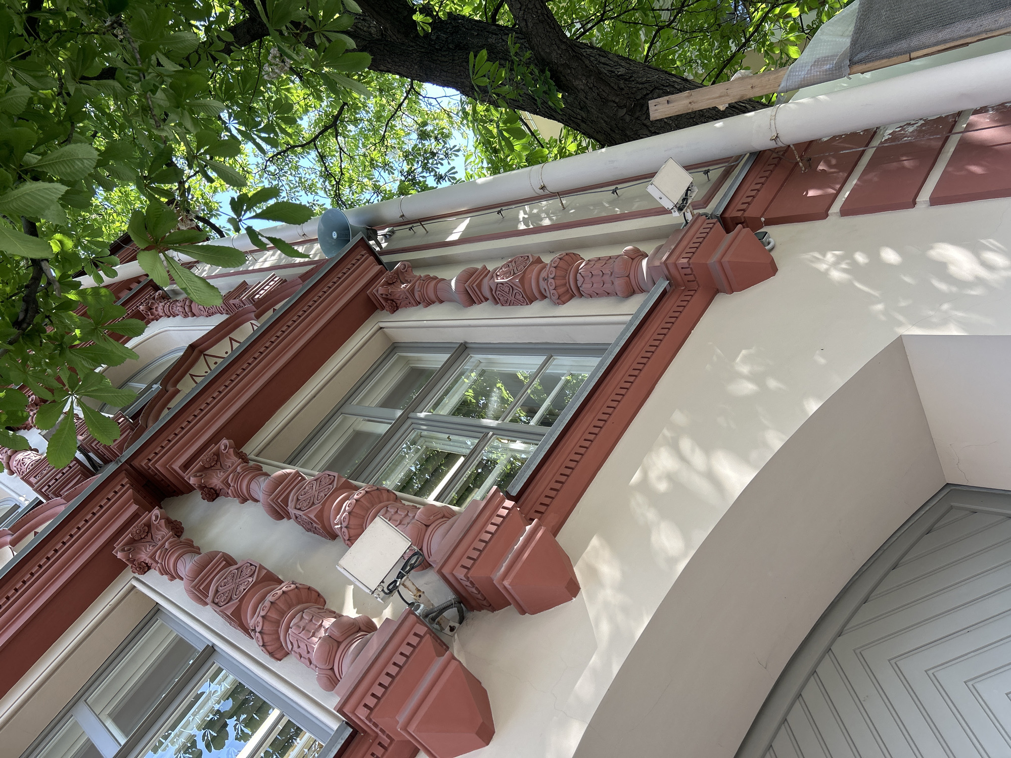 A low-angle shot captures the upper facade of a building under a canopy of bright green tree leaves and a clear blue sky. The building features a cream-colored wall adorned with reddish-brown decorative elements, including ornate columns and patterned trim around large, multi-paned windows. Sunlight casts intricate, dappled shadows of the leaves onto the building's surface.
Along the top edge of the facade runs a large white pipe, likely a downspout or conduit. Mounted below this pipe are a grey, horn-shaped speaker or public address device and a rectangular white light fixture. Above the pipe on the right side, some crude wooden planks and clear plastic sheeting are visible, suggesting temporary work or covering.
The lush green foliage of a large tree, possibly a horse chestnut due to its distinct palmate leaf shape, dominates the upper left portion of the image and extends across the clear blue sky. Its sturdy trunk is also visible. The windows reflect the surrounding greenery and sky.
Part of an arched entrance or doorway with light blue/grey diagonal paneled detailing is visible on the lower right. The scene is brightly lit by natural sunlight, indicating daytime with clear weather. There is no discernible activity taking place, nor is any text visible in the image. The architectural style suggests an older building, possibly European.