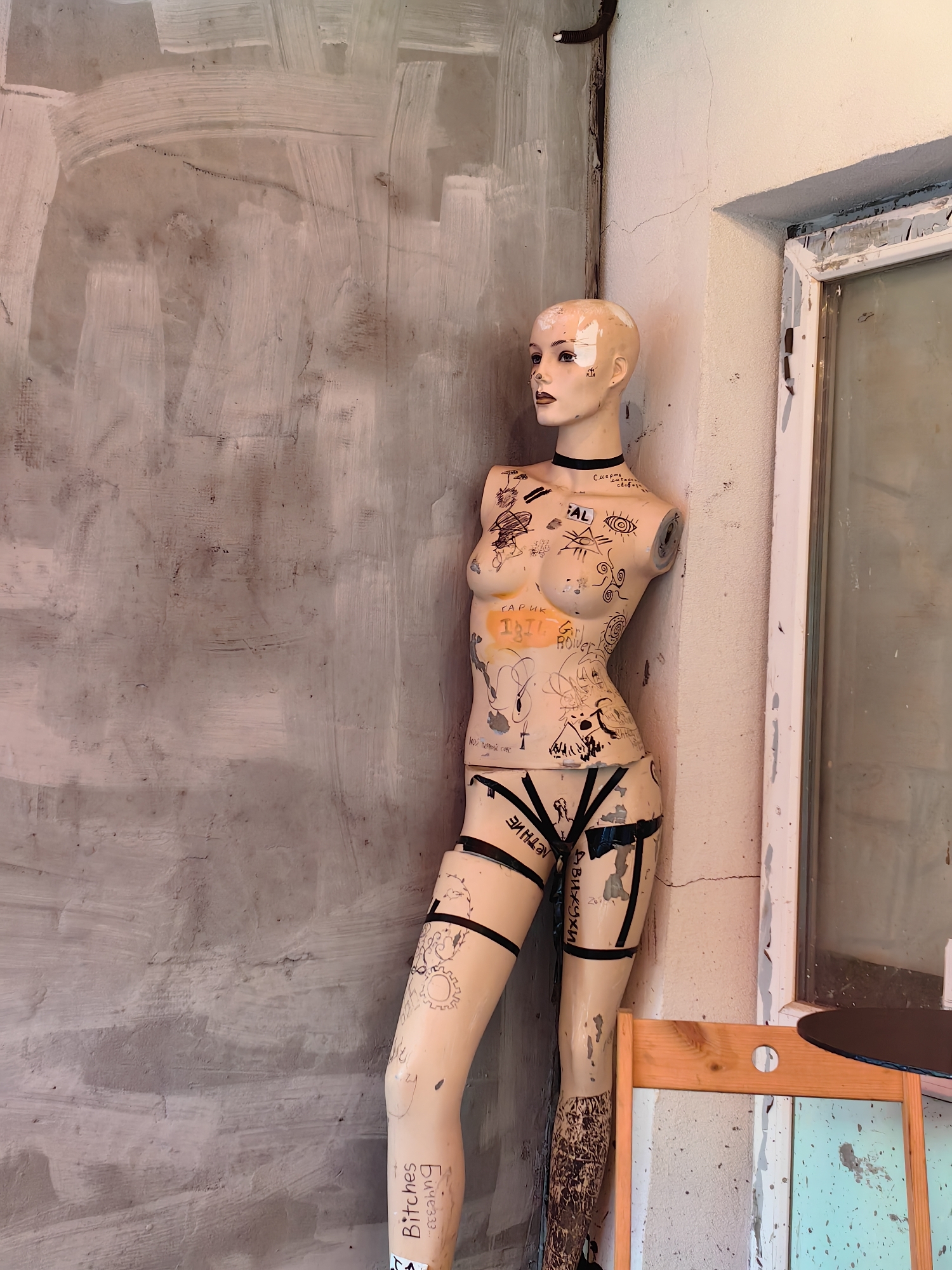 The image depicts a female mannequin, seemingly abandoned or intentionally placed in a corner of what appears to be an outdoor urban setting. The mannequin is positioned leaning against a textured, weathered wall, with a portion of a white-painted structure and a window frame visible to its right. The mannequin itself is adorned with various graffiti and markings.
The wall behind the mannequin is rough and appears to have been painted with broad, uneven strokes of a light color, possibly a distressed plaster or concrete finish. There are visible cracks and imperfections on this surface. To the right, a white-painted corner and window frame suggest an adjacent building or structure. The window appears to be a single pane, with a slightly obscured view of what might be another area or a wall.
The mannequin is unclothed from the chest up, with its arms missing. Its skin tone is pale, and it has makeup applied to its face, including prominent eyes and lips. Black bands are visible around its neck and as straps across its torso and thighs, resembling a harness or lingerie. The mannequin's body is covered in black and colored graffiti, including drawings of eyes, abstract shapes, text, and what appear to be floral motifs on its legs. Some of the markings are in Russian, such as "Смерть" (Smert' - Death) and "Girl Power".
In the foreground, to the lower right of the mannequin, a simple wooden chair with a light blue seat is visible. Part of a dark circular tabletop from what seems to be an outdoor table is also present. The lighting suggests it is daytime, with shadows indicating a bright light source.
The context provided places this scene in Ulyanovsk, Russia. This specific location is likely an outdoor area of a commercial establishment, a public space, or perhaps a forgotten corner of the city where such a display might be found. The overall impression is one of urban decay and artistic intervention, where a discarded mannequin becomes a canvas for graffiti.