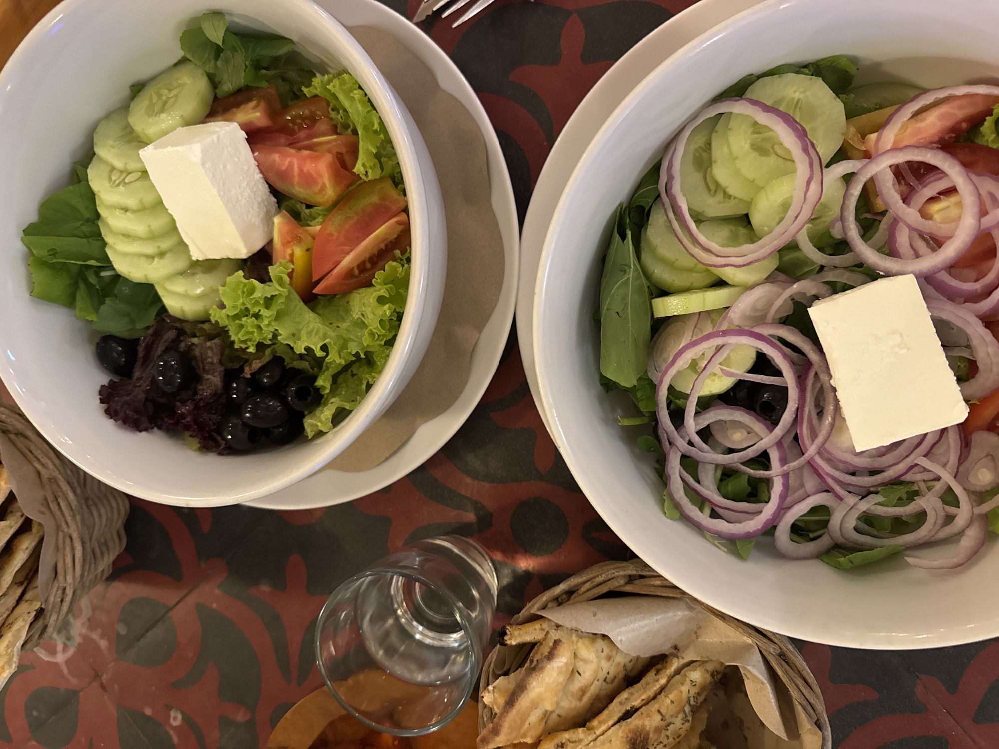 Two bowls of salad, containing lettuce, cucumber, tomato, red onion, feta cheese, and olives, are arranged on a surface with a red and black pattern. One bowl also contains bread and an empty glass. The location is Ungasan, Indonesia.