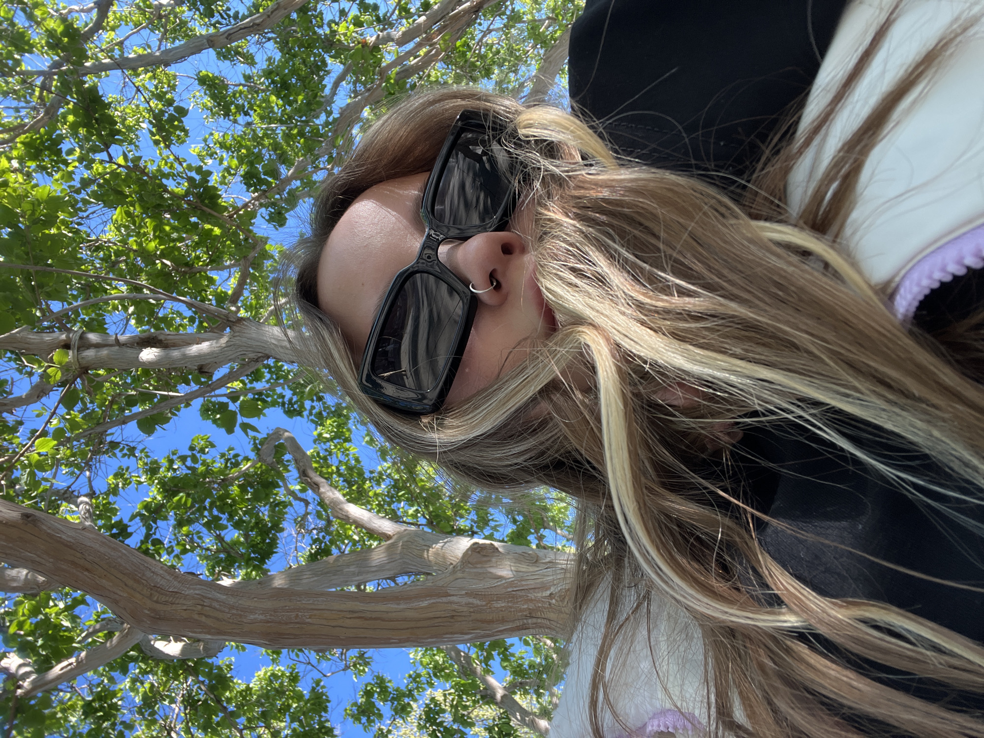 A woman is viewed from a low angle, presumably by someone lying beneath her, capturing her face, sunglasses, hair, and torso against a backdrop of tree branches, leaves, and a blue sky. She has a nose ring. Her hair is styled with blonde highlights. She appears to be wearing a dark-colored top with a white collar trim and purple accents. The image does not offer information that it was taken in Krasnodar, Russia.