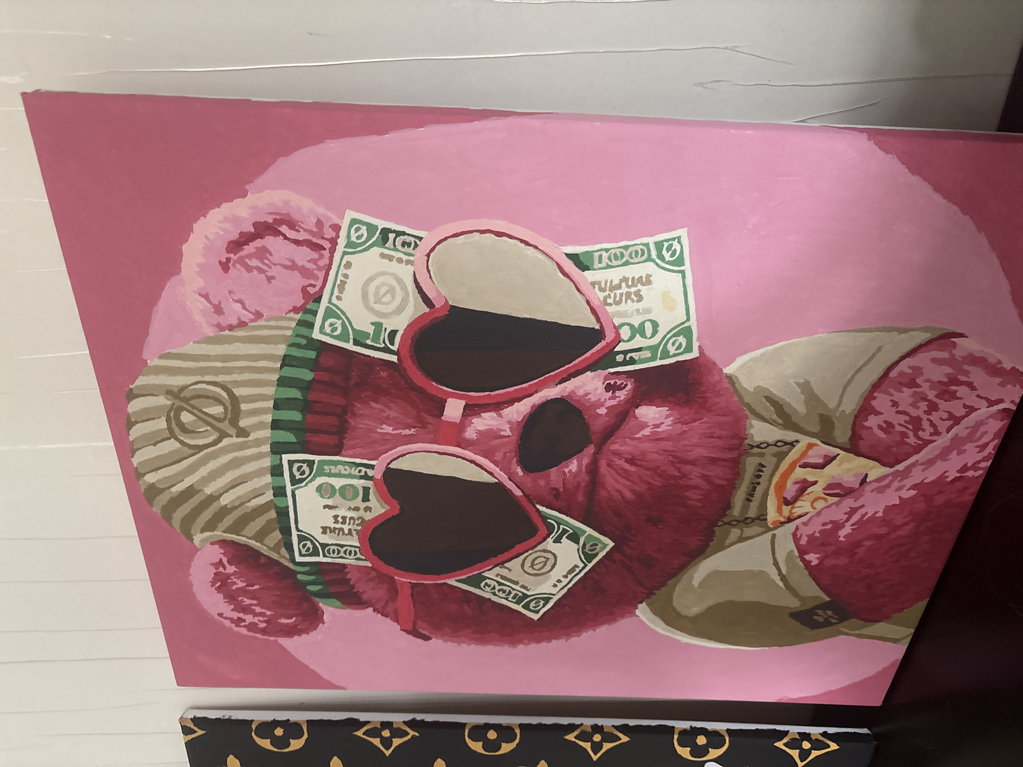 The image displays a vibrant painting featuring a large, fluffy, magenta-colored teddy bear. The bear is depicted wearing prominent heart-shaped sunglasses with pink frames and dark, partially reflective lenses. Numerous stylized dollar bills are scattered around the bear's head and neck. These bills are green and marked with "100" and the partial text "FUTURE CURS," some appearing mirrored.
On the bear's left side, it sports a striped accessory, possibly a beanie or hat, in shades of brown and green, adorned with a distinct gold circular symbol. On its right, a beige or khaki-colored boot or shoe is visible, with straps and a small dark square detail. The painting's background consists of two shades of pink, with a lighter, circular halo behind the bear's head, creating a soft aesthetic.
The artwork, rendered on canvas, is displayed leaning against a white surface, suggesting an indoor setting. A thin silver wire is visible at the top of the frame. A fragment of another canvas, featuring a black background with a repeating gold pattern resembling a luxury brand monogram, is partially visible at the bottom of the frame. This static scene evokes themes of playful luxury and extravagance. No specific activity or event is taking place, and the time of day or weather is not discernible. The location context provided is Ореховка, Russia.