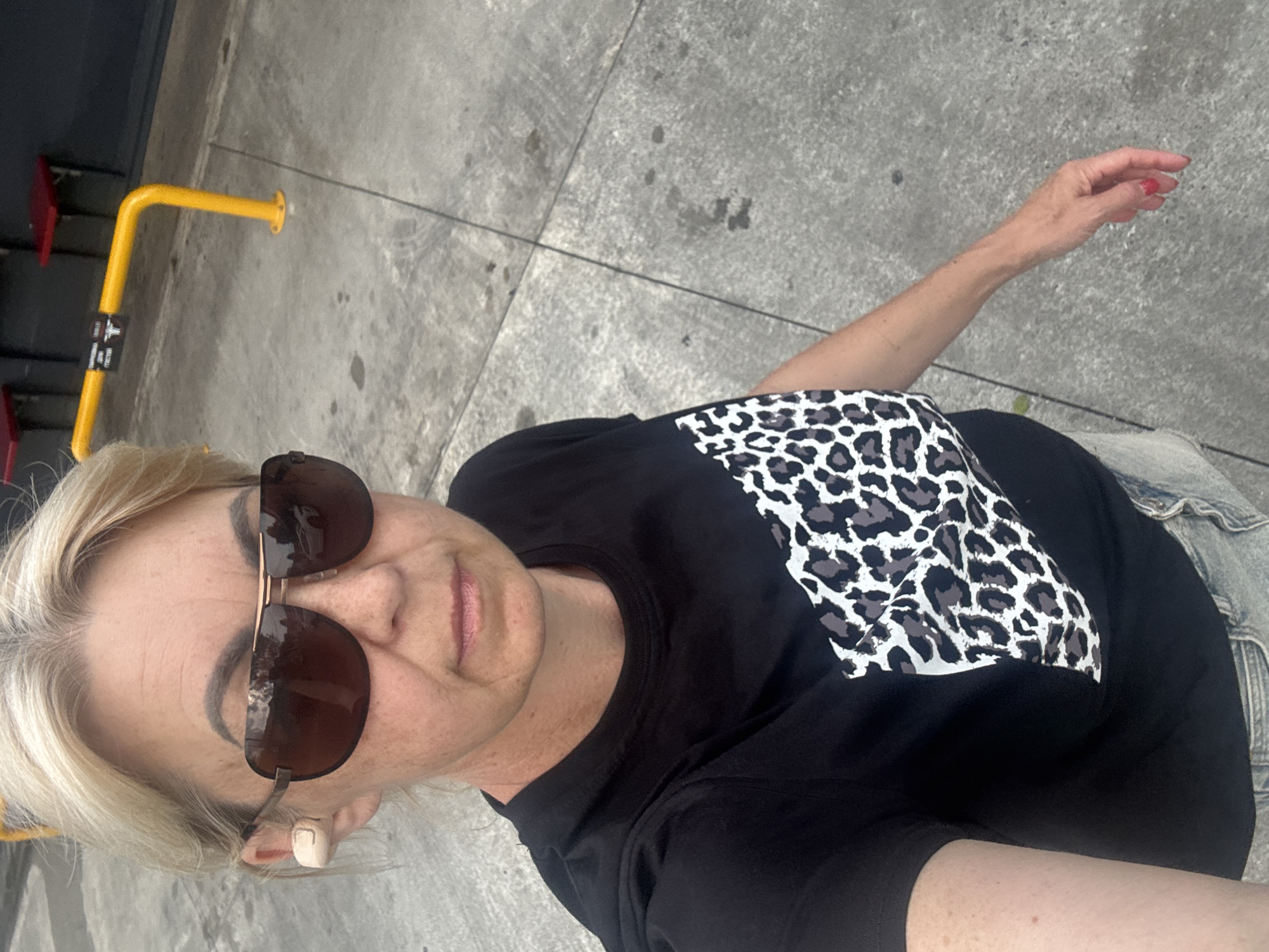 A blonde woman wearing aviator sunglasses and a black t-shirt with a leopard print design is standing outdoors on a concrete surface. A beige wireless earbud is visible in her left ear. Her fingernails are painted red. In the background, a yellow metal railing and red seats are visible. The location is not the city of Sochi, Russia, as there are no contextual clues to suggest this location.