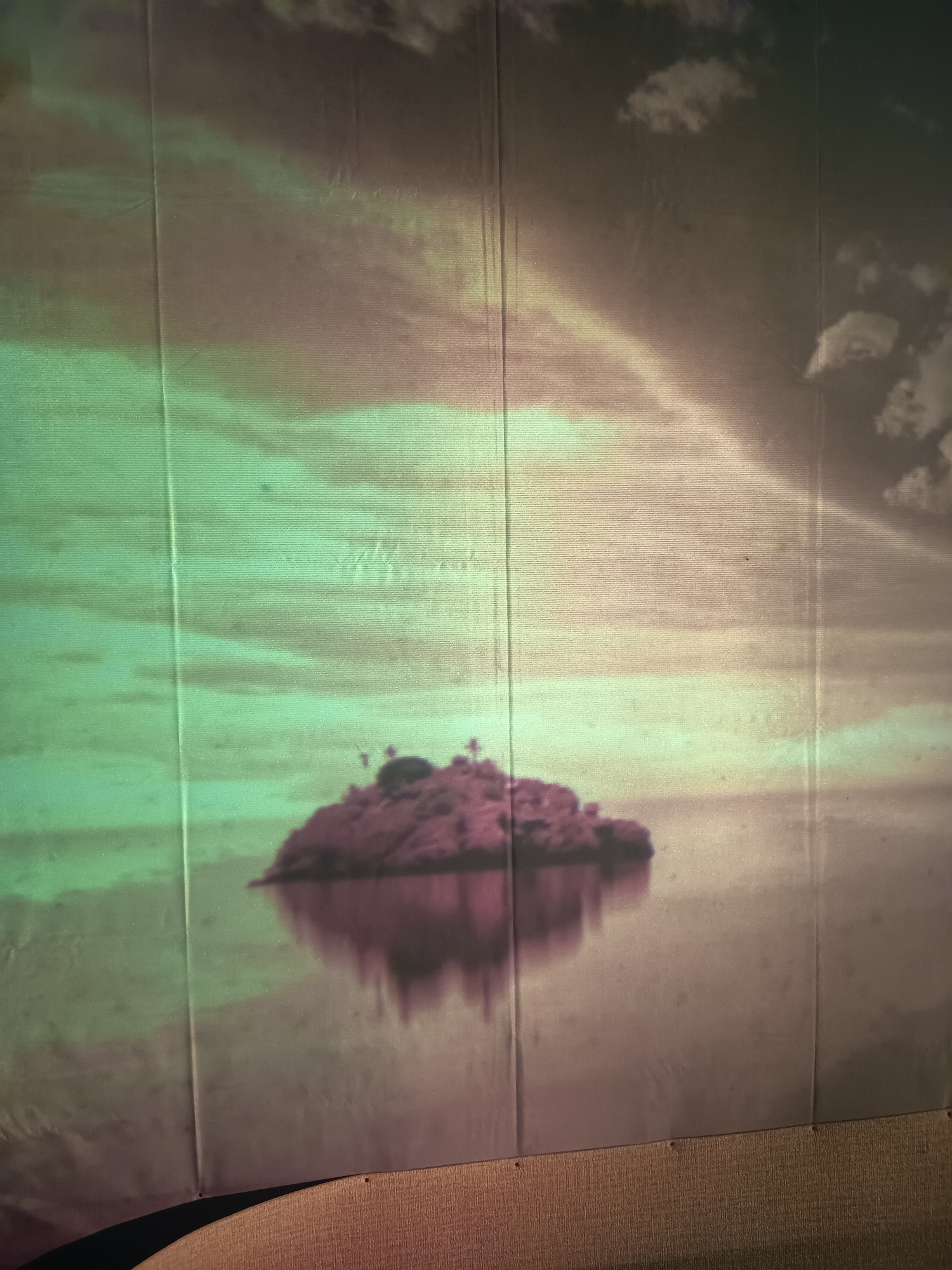 This is a projected image of a small island with palm trees silhouetted against a cloudy sky. The island is reflected in the water below. The image is projected onto a textured surface, possibly a fabric screen, which shows vertical lines from its construction and some minor damage or dirt marks. The lighting appears to be a green and pinkish hue, creating an atmospheric effect. The bottom of the image shows a wooden surface and a dark, curved object. There are no people or animals visible. The scene is a natural landscape and does not depict any specific location within the city of Kalipenggung, Indonesia.