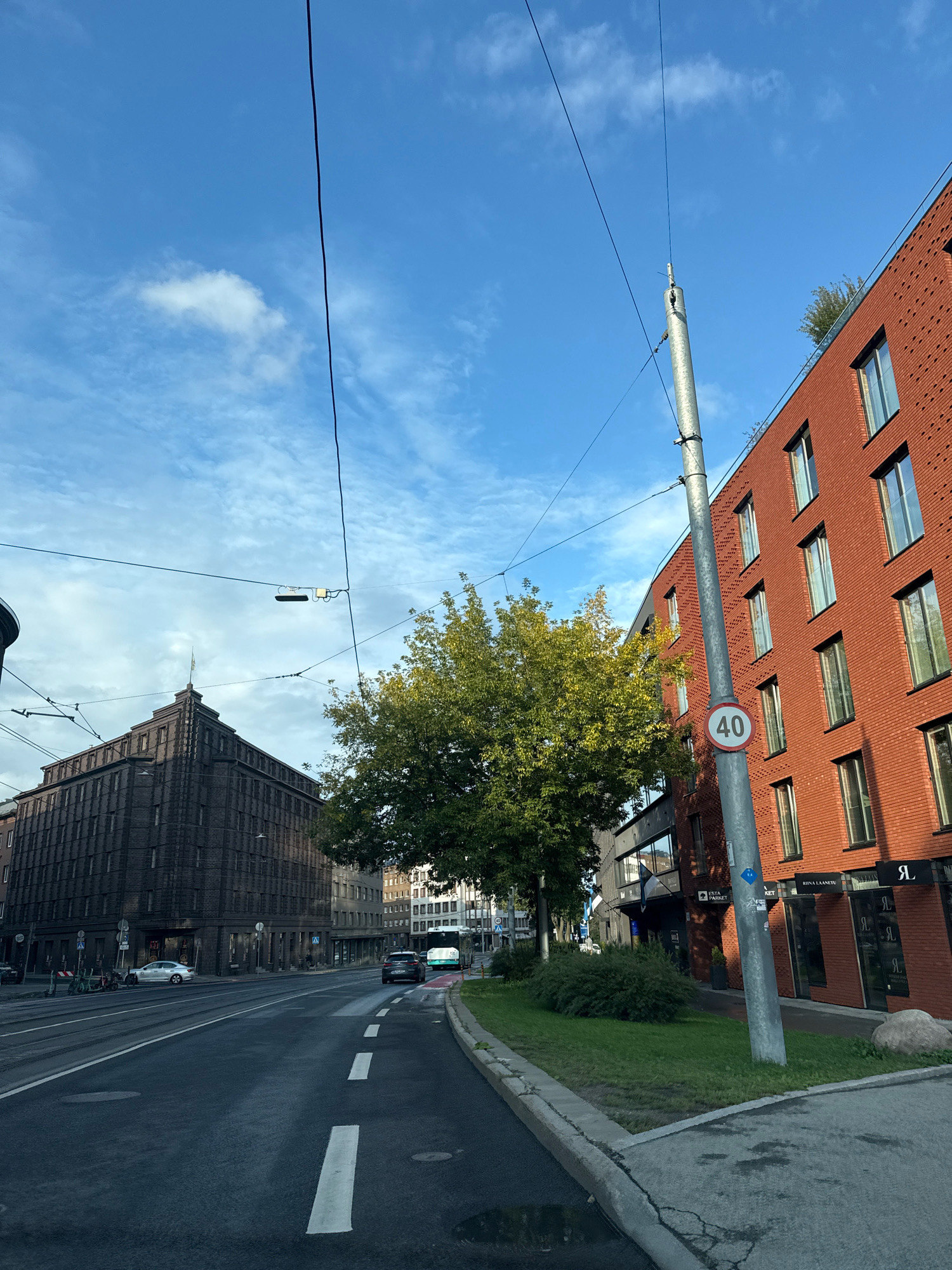 The image captures a view of a street in Tallinn, Estonia, with a prominent red brick building on the right and a darker, more ornate building on the left. A street pole with a speed limit sign showing "40" is positioned between the two buildings. Overhead, power lines and trolley lines crisscross the bright blue sky with scattered clouds.
On the right, the modern red brick building features multiple floors with large windows, some reflecting the sky. Below, storefronts with signage like "Riina Laanetu" and a stylized "Я" are visible. A large rock sits on the grassy area in front of this building.
The building on the left is characterized by its dark brick facade, intricate architectural details, and a distinct stepped roofline culminating in a flagpole. In front of this building, a street with tram tracks runs, and a silver car is parked. Bicycles are also visible near the curb.
In the background, more buildings of varying architectural styles line the street, including a bus with "20" visible on its side. A green tree with lush foliage stands between the two main buildings. The road surface appears wet in places, suggesting recent rain. The overall scene depicts a typical urban streetscape in Tallinn.