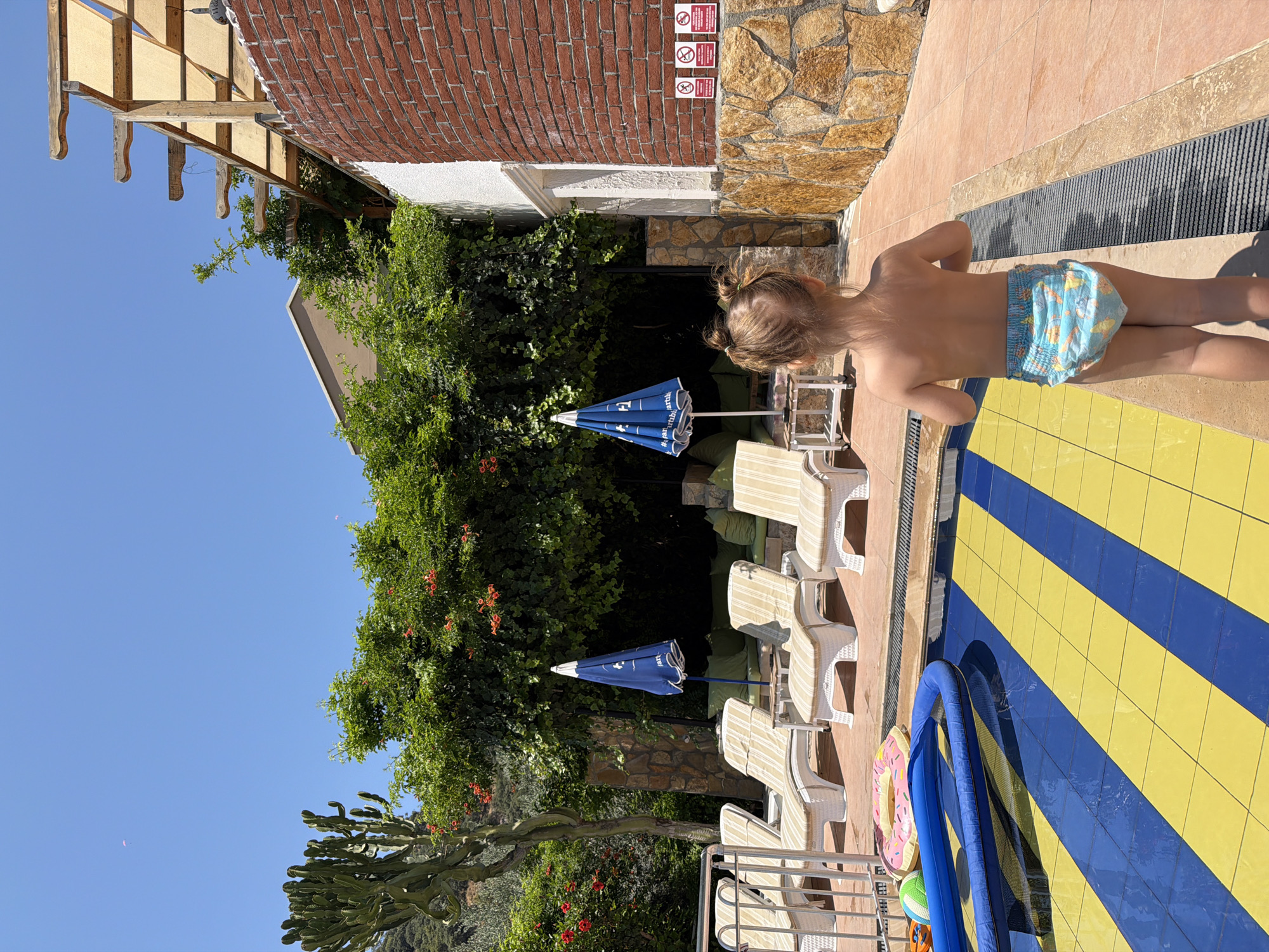 A young child, viewed from behind, stands on a tiled pool deck near the edge of a swimming pool. The child is wearing a blue swim diaper with a cartoon print. The pool deck is a vibrant pattern of yellow and blue tiles. To the left of the child, several white lounge chairs are arranged, some with striped cushions, next to a blue umbrella. Lush green foliage, including a cactus and trees with red flowers, surrounds the lounge chairs. In the background, a brick building and a stone wall are visible. The sky is a clear, bright blue, suggesting a sunny day. A wooden awning with a tan fabric covering extends from the building. Two small red signs with white symbols are mounted on the brick wall. The overall scene depicts a relaxed, summery atmosphere at a poolside resort, likely during the daytime.