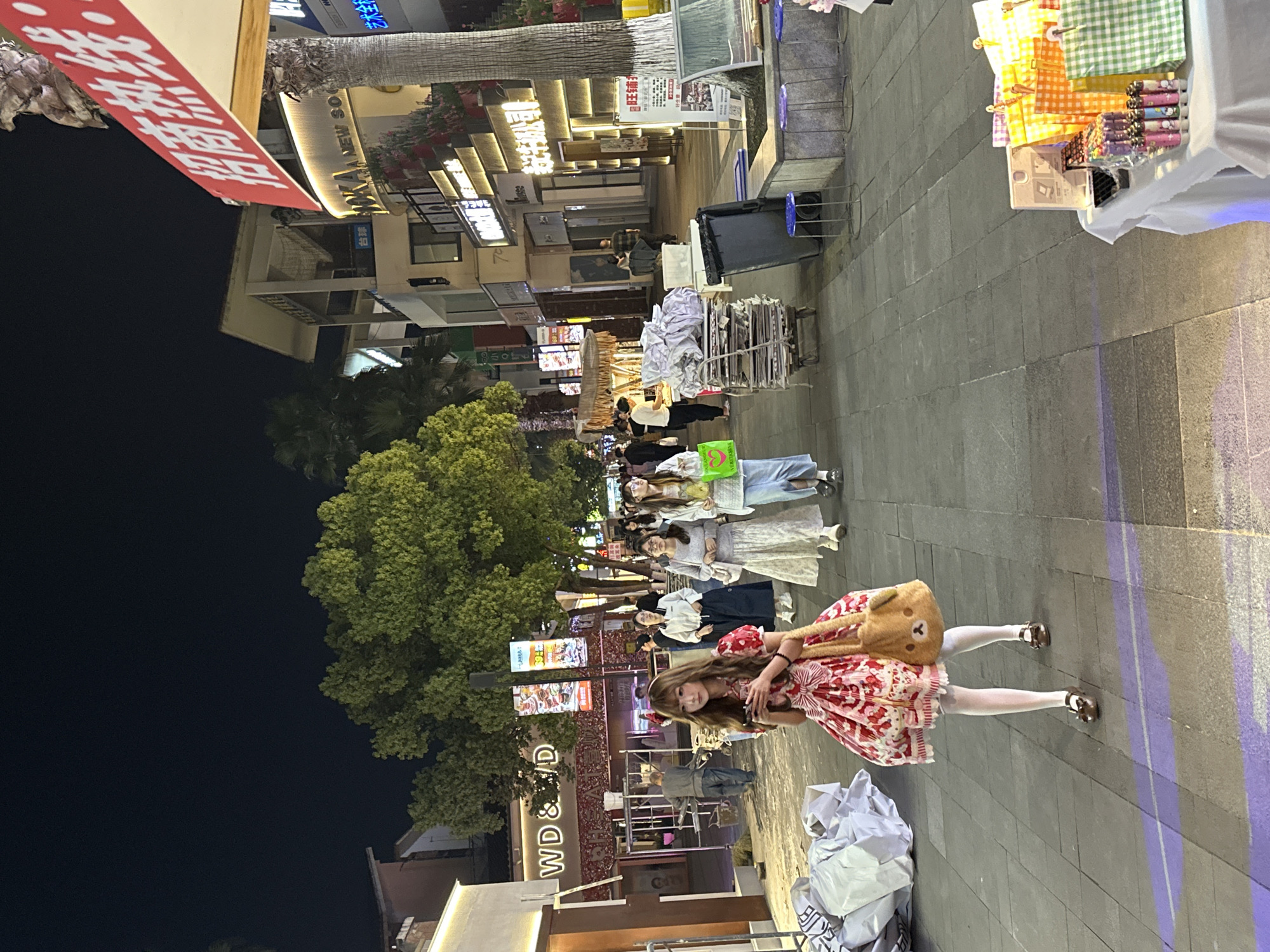 The image shows a group of people on a street in what appears to be a commercial area. The street has several shops and businesses, as indicated by the signs and storefronts. At the front of the group is a young woman in a red and white dress with white tights and a bear bag, posing for the picture. Behind her, other individuals are walking along the street. The scene is set at night, with artificial lights illuminating the area. Signage suggests this is the Chongqing High-tech Zone, China.