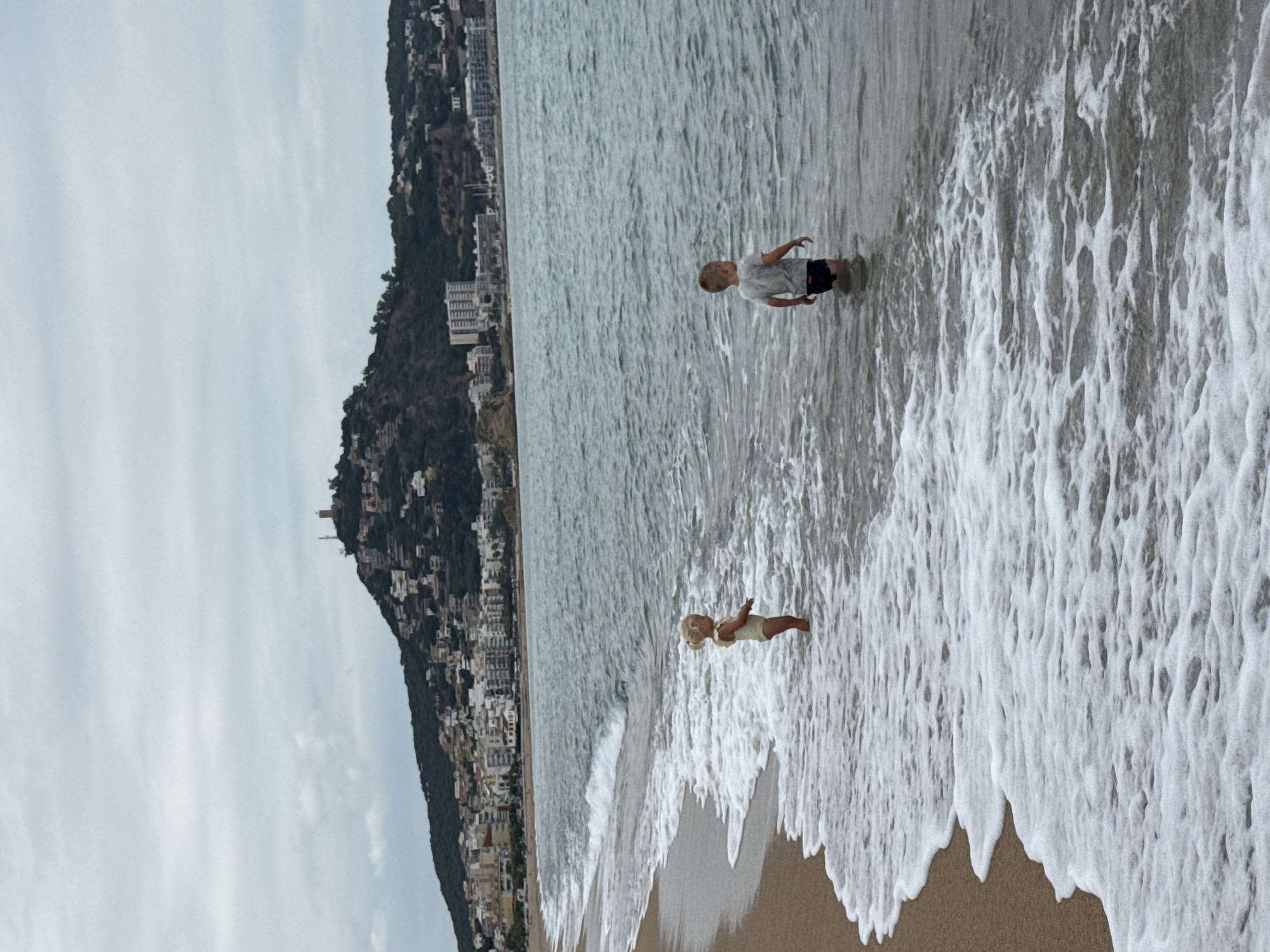 A full-frame, medium shot captures two young children wading in the gentle waves of a beach in Blanes, Spain. The girl, on the left, stands facing to the right, her blonde hair tied back, wearing a pale yellow swimsuit. The boy, on the right, stands facing away from the camera, wearing a grey t-shirt and dark shorts. He is positioned further out in the water than the girl. The sea, with its foamy white waves, is calm and extends towards a distant shoreline. Behind the water, a densely populated town sprawls across the foothills of a large, verdant hill. Buildings of various sizes and colors are visible, with a prominent tower at the summit of the hill. The sky is overcast with a soft, grey light. The foreground is dominated by the sandy beach and the encroaching waves, creating a sense of gentle motion and a tranquil atmosphere.