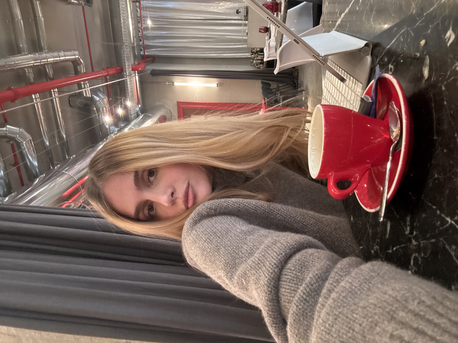 A female individual with long blonde hair and a grey turtleneck sweater is seated at a dark, veined marble table, looking directly at the camera. On the table, a red ceramic coffee cup with a white interior rests on a matching red saucer, accompanied by a silver spoon and a small blue and white packet. Behind the cup, an open light-colored laptop or tablet with a keyboard case is angled. The background depicts an indoor commercial establishment, featuring dark grey curtains on the left. Further back, a dining area with white chairs and dark tables, some adorned with red glasses or objects, is visible. A red door frame is prominent in the mid-background. The ceiling displays an industrial design with exposed silver metallic ventilation ducts and red pipes, illuminated by multiple recessed spotlights and a vertical tube light fixture mounted on a grey wall. The scene is set within a commercial space in the city of Unknown, Unknown.