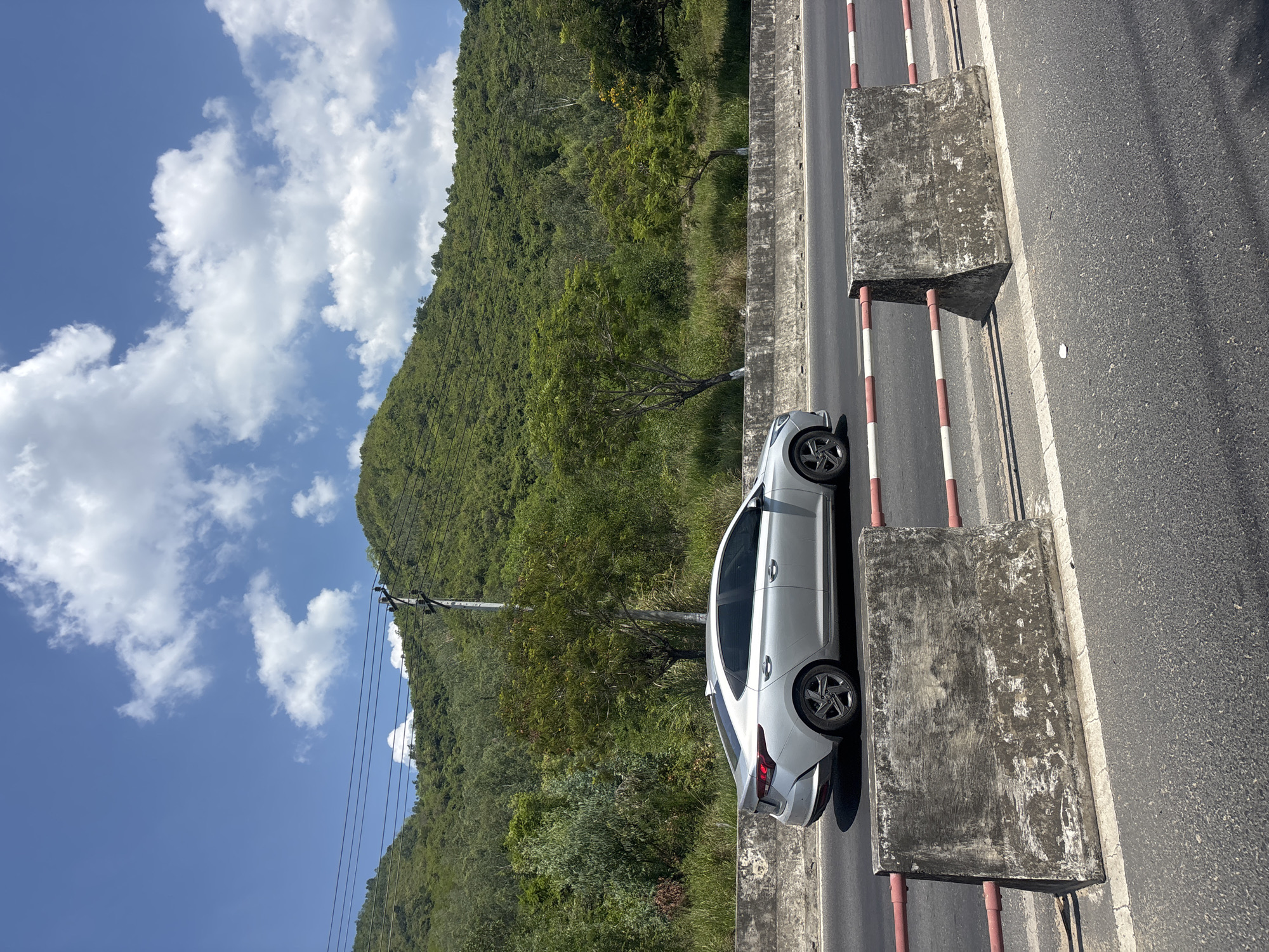 In a natural, outdoor environment, a silver sedan is captured in profile, driving on an asphalt road. The car is positioned mid-frame, moving from left to right. The road is lined with concrete barriers. Behind the road, a lush green mountainside is covered in dense vegetation and trees, with power lines and a utility pole visible in the upper left quadrant. The sky above is a bright blue, dotted with fluffy white clouds. The scene appears to be a sunny day.