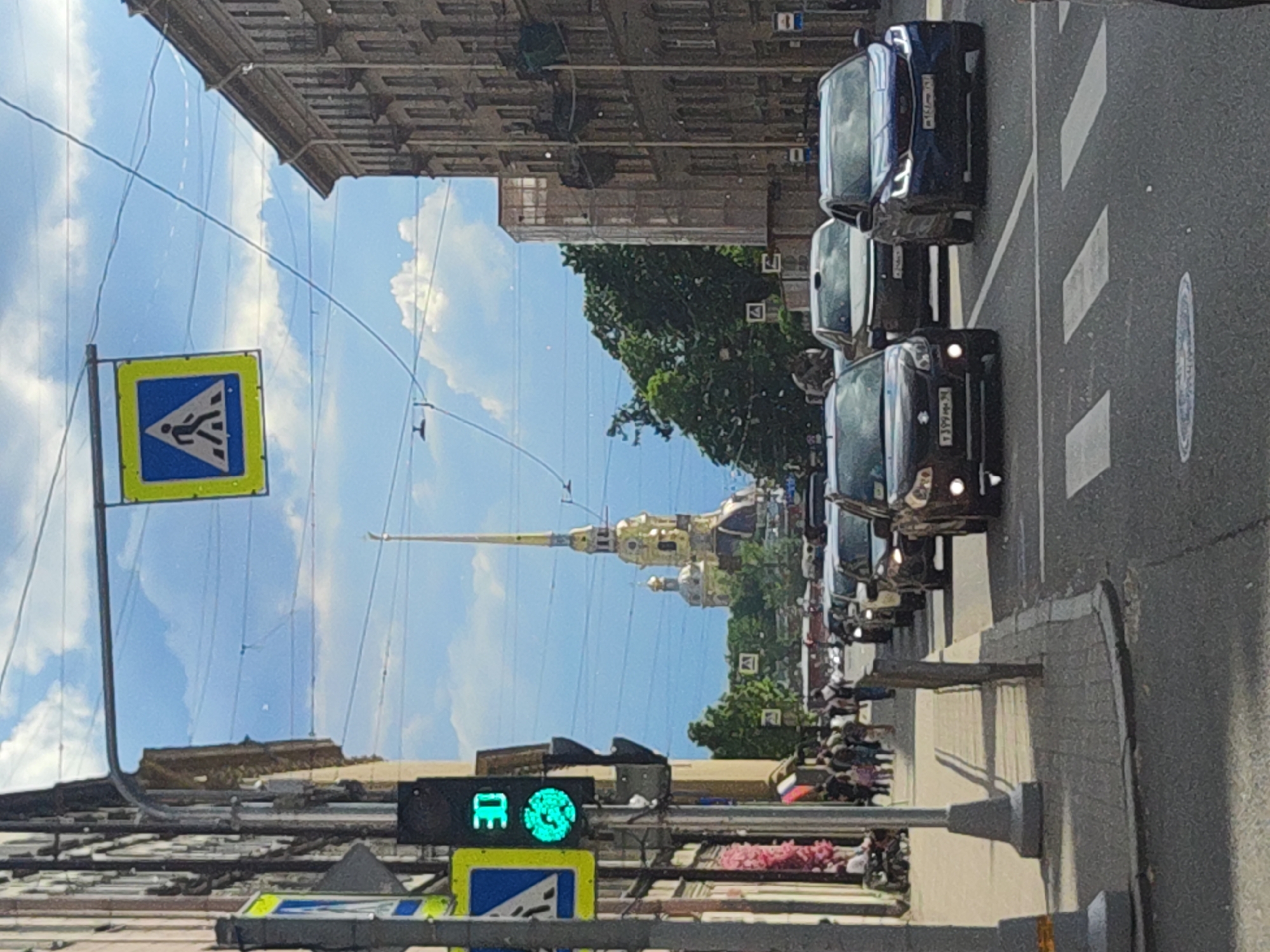 The image captures a street scene in Saint Petersburg, Russia, likely on a sunny day with scattered clouds. In the foreground, a traffic light displays a green signal and a digital readout of "30," indicating a speed limit. Pedestrian crossing signs are visible on either side of the street. Several cars are proceeding down the road, some stopped at the pedestrian crossing. The architecture of the buildings lining the street is characteristic of Saint Petersburg, with ornate facades and balconies. In the distance, the spire of the Peter and Paul Cathedral, a prominent landmark, rises above the cityscape, its golden dome glinting in the sunlight. The street is paved with cobblestones, and trees line the sidewalks. People are seen walking on the sidewalk and near the buildings, engaged in various activities. The overall atmosphere is one of a bustling urban environment with a blend of historical and modern elements.