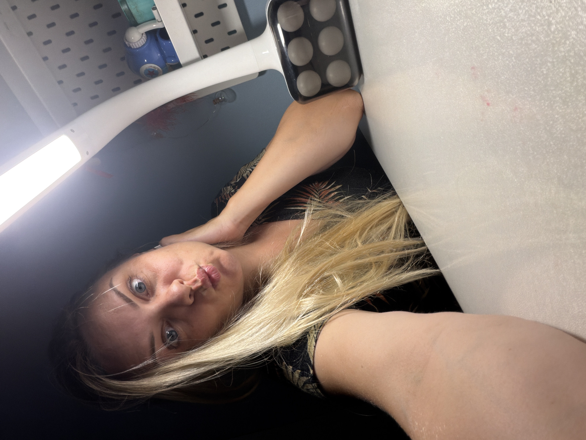 A woman with blonde hair and blue eyes is taking a selfie at a desk in Votkinsk, Russia. She is wearing a black shirt with a tropical print. A desk lamp is positioned to her right, and a pegboard with various items is visible in the background. The lighting in the scene is dim, with the desk lamp providing the primary light source. The woman is puckering her lips and touching her neck with her left hand. The desk surface is white and appears to have some pink marks on it.