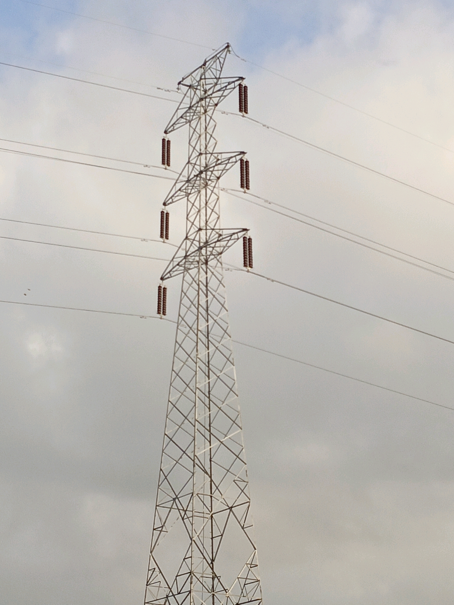 The image shows a tall, lattice-style electricity pylon set against a cloudy sky. The pylon is made of metal and has several crossbars supporting thick power lines. Three sets of insulators, each consisting of multiple brown cylindrical components, are visible hanging from the crossbars, connecting them to the power lines. The power lines stretch diagonally across the frame. There are no people or animals depicted in the image. The environment appears to be an open area, possibly rural or semi-urban, given the presence of such infrastructure. The lighting suggests it is daytime, possibly overcast. Without additional context, the specific location within Bulukumba, Indonesia, cannot be determined from the image alone, as such pylons are common in many areas.