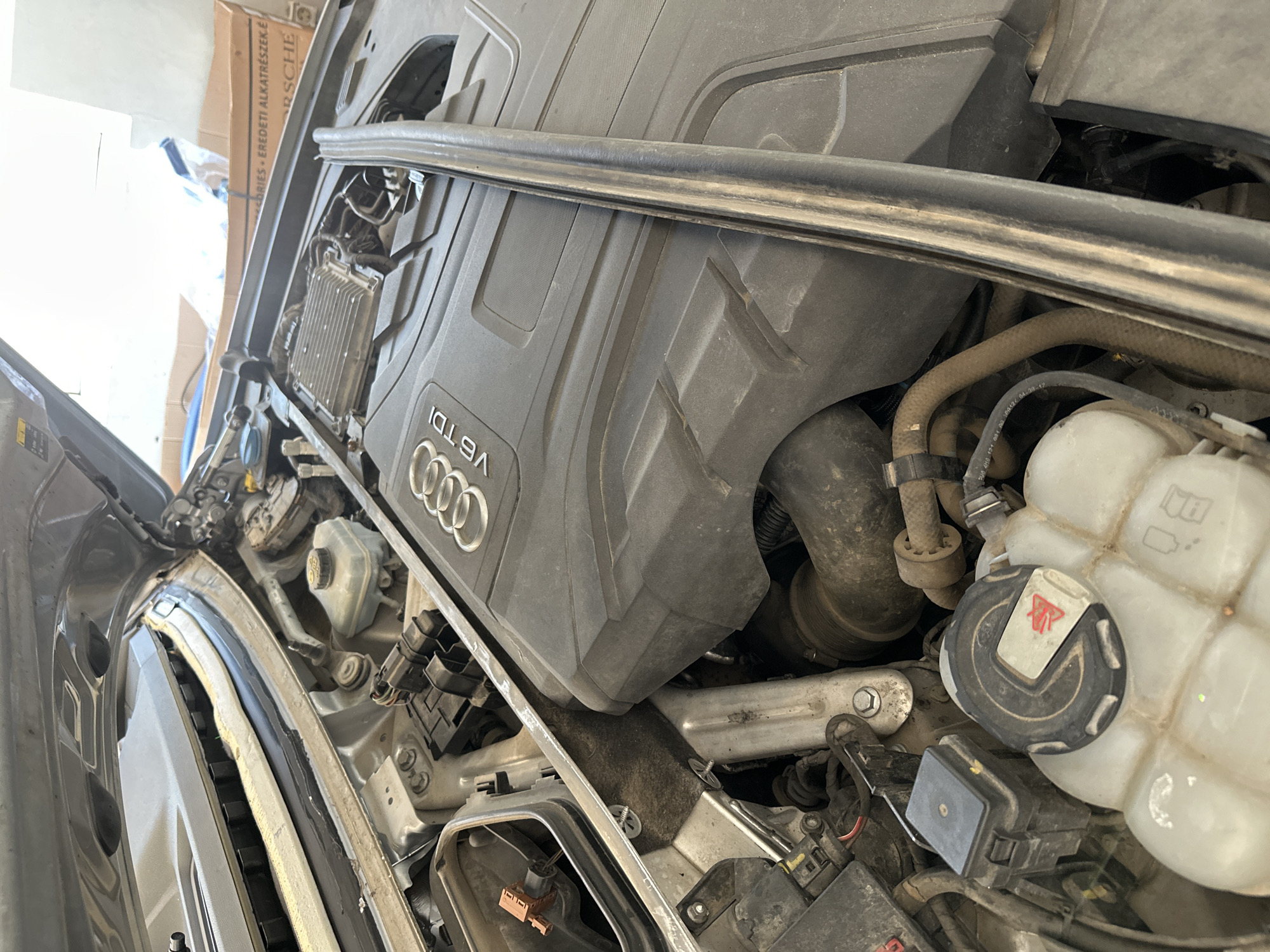 The image shows the engine compartment of a car, likely an Audi, with the hood open. The engine is partially covered by a black plastic engine cover with "V6 TDI" branding. Various engine components, hoses, and wiring are visible. A silver metal bar runs vertically through the center of the image, obscuring part of the engine. In the foreground, a translucent plastic coolant reservoir with a black cap is visible. The overall environment appears to be a workshop or garage, suggested by the presence of car parts and a cardboard box in the background. The scene is captured from a slightly elevated perspective, looking down into the engine bay.