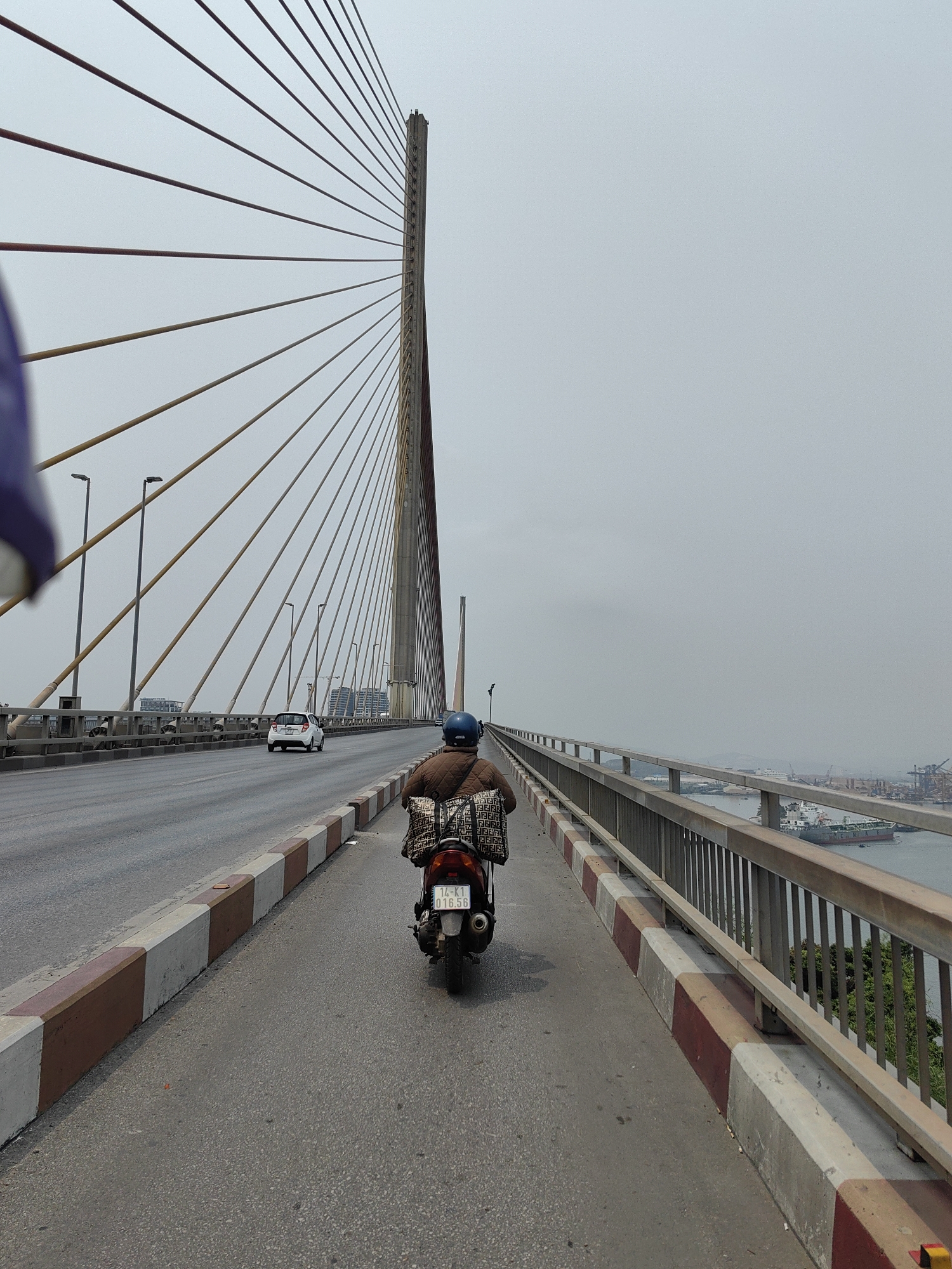 The image depicts a person riding a scooter on Bai Chay Bridge in Hong Gai Ward, Vietnam. The bridge is a cable-stayed structure with a prominent tower and numerous cables extending outwards. The scooter rider is wearing a brown jacket and a blue helmet, with a patterned bag on the back of the scooter. The scooter's license plate is visible. A white car is present on the bridge's main lane. The scene is set on an overcast day, with the sky appearing hazy. On the right side of the bridge, a metal railing overlooks a body of water with ships and port infrastructure visible in the distance.