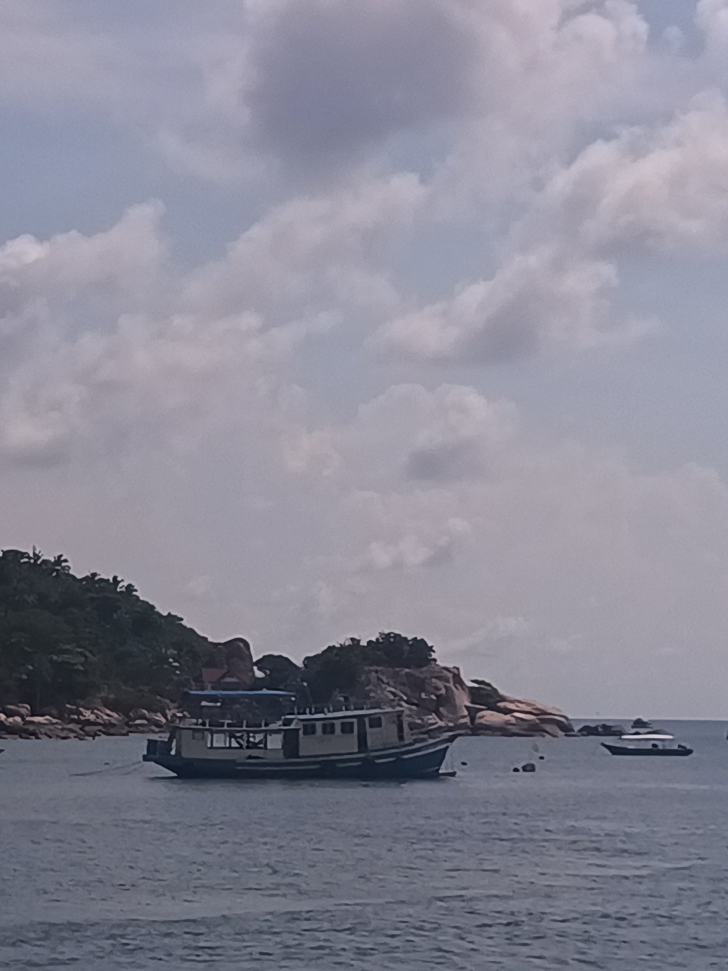 The image depicts a marine scene in Tambon Ko Tao, Thailand. A medium-sized boat is anchored in the foreground, with its hull displaying a mix of light and dark colors. In the background, a landmass features lush green vegetation and rock formations. Another, smaller boat is visible further in the distance. The sky is overcast, with scattered clouds. The water appears calm, with minor undulations.