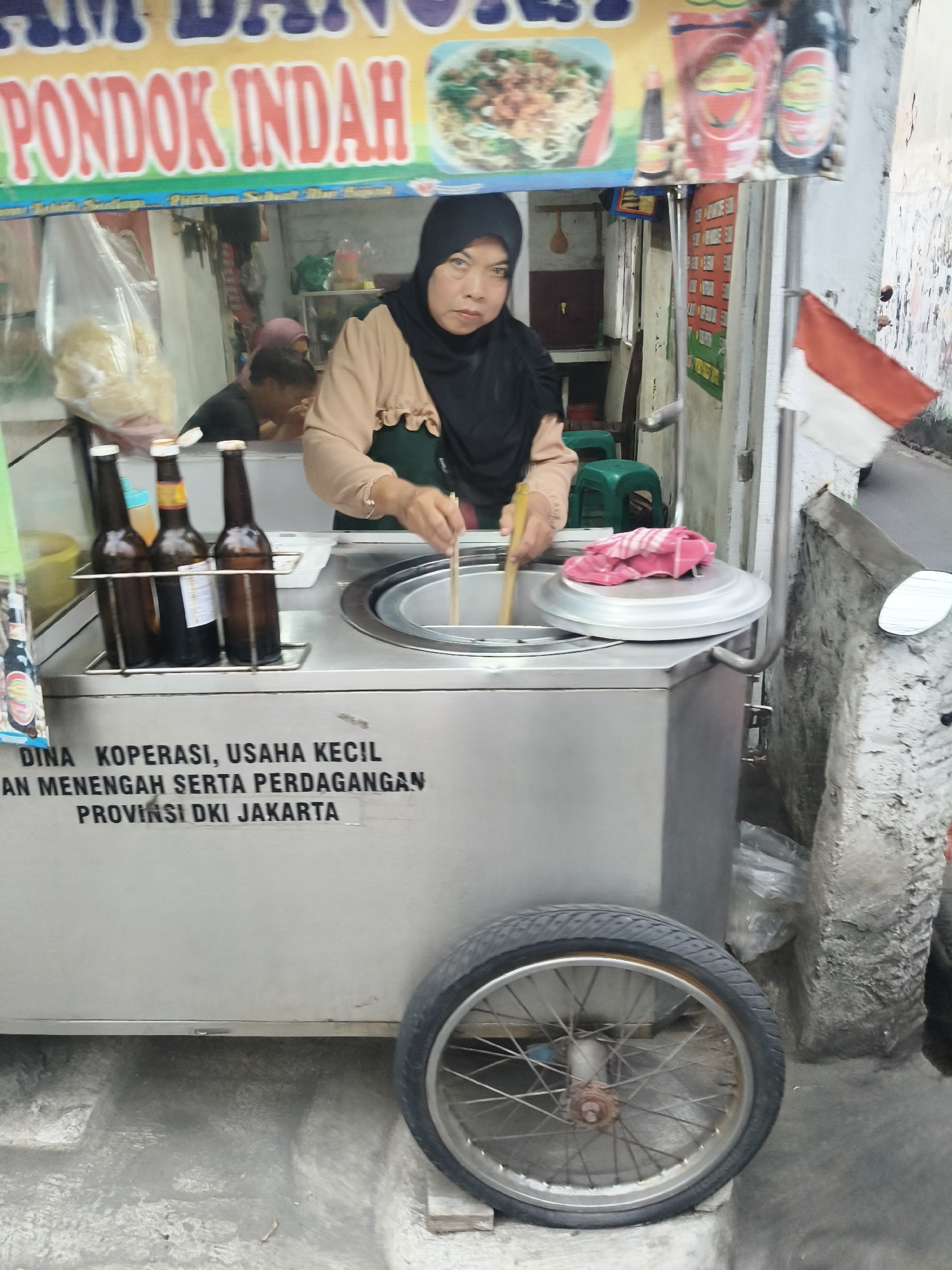 A middle-aged Indonesian woman wearing a black hijab and a beige top is preparing food in a food cart on a street in South Tangerang, Indonesia. The food cart has a large metal container with a lid, and the woman is holding two chopsticks, stirring something inside the container. There are three bottles of what appears to be soy sauce or similar condiments on a rack in front of the container. A red and white Indonesian flag is visible on the right side of the cart. The background shows a busy street scene with other food stalls and people. The overall impression is that of a typical street food vendor in an Indonesian city.