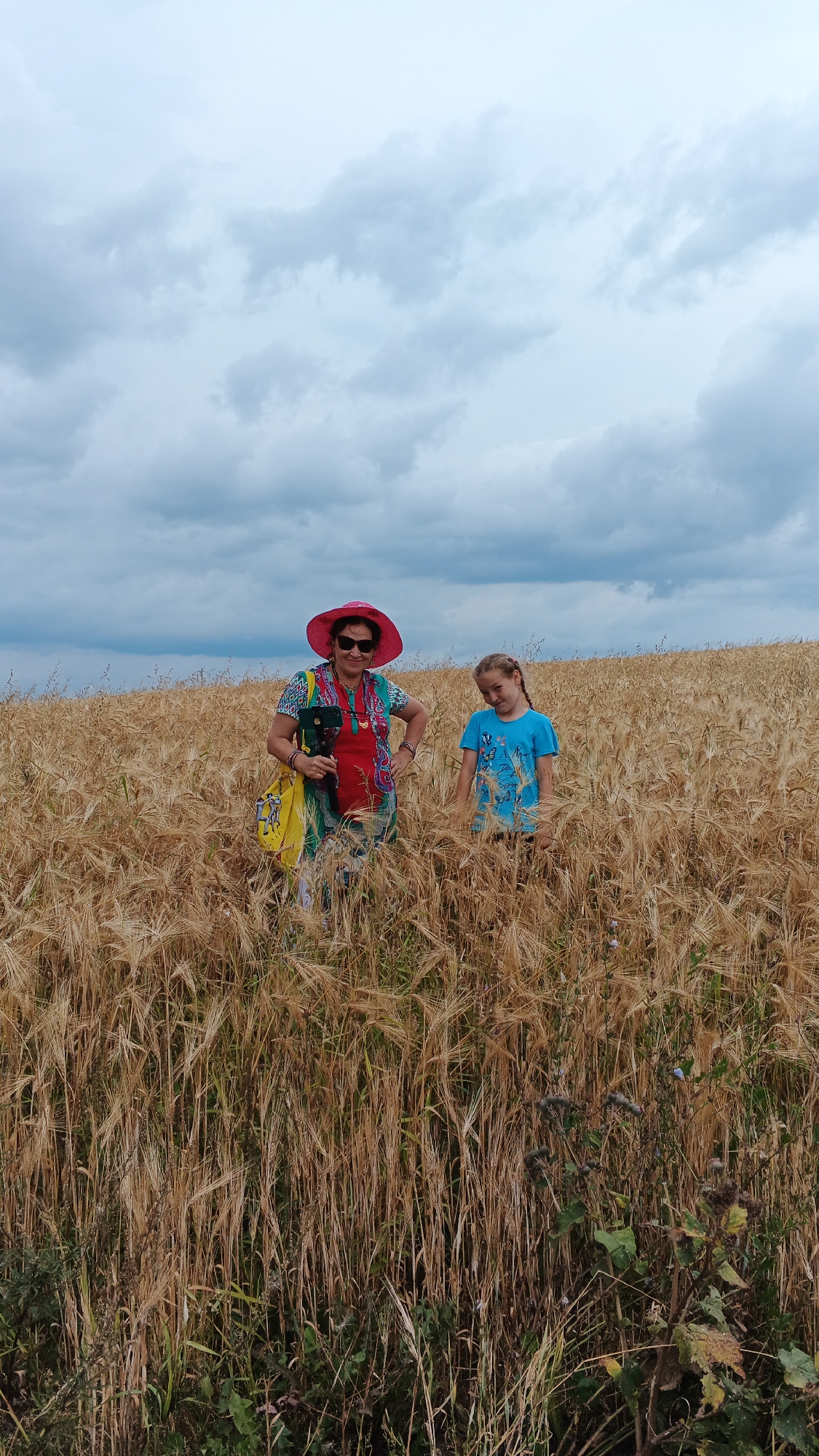 The image depicts two individuals, an adult and a child, standing in a field of dry, golden wheat under a cloudy sky. The adult, positioned on the left, wears a vibrant red and green patterned dress, a bright pink wide-brimmed hat, sunglasses, and several bracelets. They are holding a dark-colored object, possibly a small instrument or camera, and have a yellow bag slung over their shoulder. The child, on the right, is wearing a blue t-shirt with a butterfly design and has braided blonde hair. They are looking towards the camera with a slight tilt of their head. The wheat stalks are tall and densely packed, obscuring much of the lower bodies of the individuals. The background shows a consistent expanse of wheat stretching towards the horizon, with a dramatic, overcast sky overhead. The location is described as the city of Podgornoye, Russia.