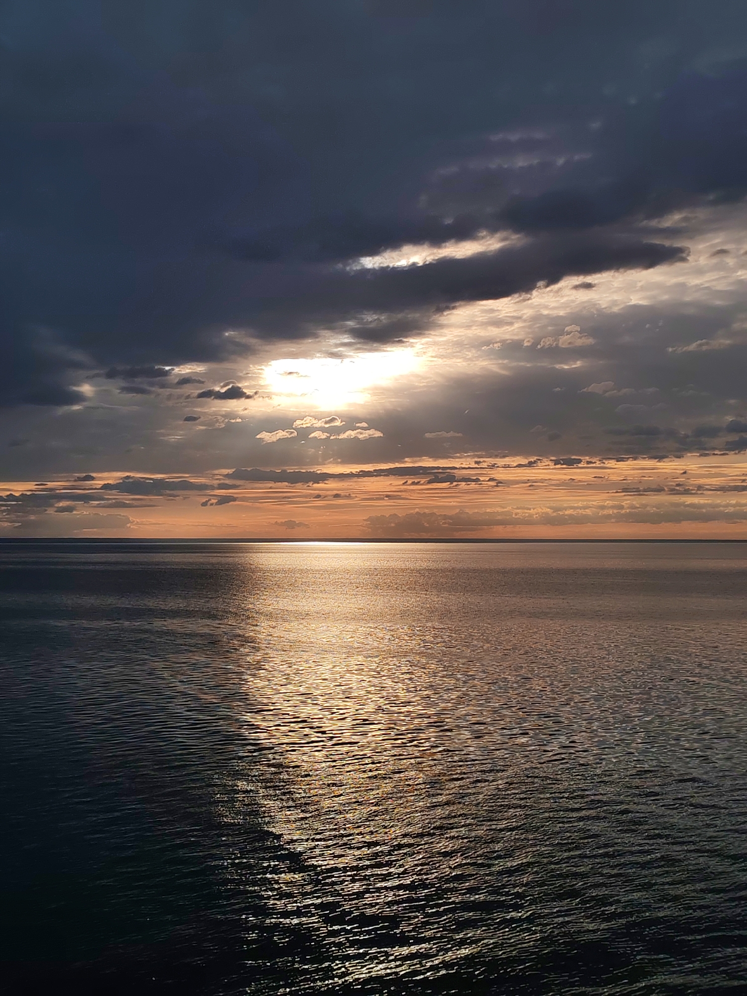 The image depicts a body of water, possibly a sea or a large lake, under a dramatic sky. The sky is filled with dark, heavy clouds, with a patch of bright sunlight breaking through. The light reflects off the water, creating a shimmering path towards the viewer. The horizon is visible in the distance. The location is not discernible from the image; no landmarks or other clues indicating Rusanovo, Russia, are present. The scene is devoid of people or other subjects. The only actions are the natural phenomena of light reflecting and water rippling.