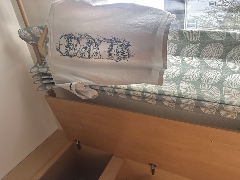 The image displays a white short-sleeved t-shirt, identified as being in a domestic interior in the city of Galway, Ireland. The t-shirt is suspended from a light-colored wooden hanger, which rests on a matching wooden curtain rod. The back of the t-shirt features a vertical graphic illustration comprising four distinct, stylized male heads. From top to bottom, text is associated with each head: "SEE NO EVIL," "HEAR NO EVIL," "SPEAK NO EVIL," and "KNOW NO EVIL." The drawing style is linear and stark.
To the left of the t-shirt, a light-toned wooden wardrobe or cupboard stands with its right door slightly ajar, revealing interior hinges and a partial view into its contents. The wall is painted a light, neutral color, and the ceiling is off-white.
To the right of the t-shirt, a sage green curtain with a repeating white leaf pattern is drawn, partially covering a window. Through the clear lower-right section of the window, the exterior of a light-colored, multi-story building, likely residential, is visible. Bare tree branches are also discernible outside, suggesting a non-summer season. The scene is illuminated by natural daylight entering from the window.