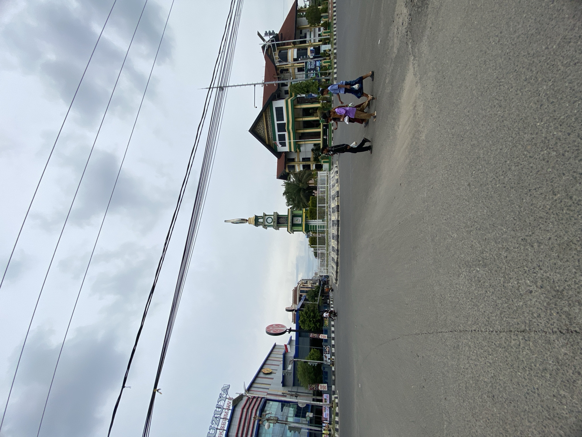 The image depicts a street scene in Lhokseumawe, Indonesia, under an overcast sky. In the foreground, an asphalt road occupies the majority of the frame, with a pedestrian crossing marked by black and white stripes. Four individuals, appearing to be children or young teenagers, are crossing the street from right to left. They are carrying bags.
Further in the background, a prominent round monument with a clock tower stands at the center of an intersection. The clock tower is green and white with gold accents and topped with a circular sculpture. To the left of the monument is a modern, multi-story building with a large sign that reads "UZUYA." This building appears to be a commercial establishment, possibly a mall or department store, with glass facades and banners displaying advertisements.
To the right of the monument, a two-story building with a dark red roof and green and yellow columns is visible. This building has a traditional architectural style. A flagpole with an Indonesian flag is positioned in front of this building. Other buildings and trees line the street in the middle ground, and overhead, numerous power lines crisscross the sky. The overall atmosphere is that of an active urban environment on a cloudy day.
