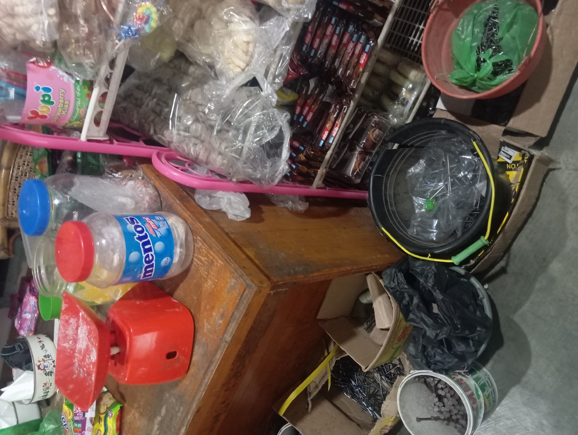This image captures a street-side stall in Rejosari, Indonesia, likely a small warung or kiosk. The scene is filled with various goods typically sold in such establishments. Dominating the foreground on the left is a bright red manual weighing scale, its pan dusted with a fine white powder, possibly flour or sugar. Beside it, a clear plastic jar filled with Mentos candies stands prominently. Behind these items, a wooden counter or table supports a collection of packaged goods and containers.
To the right, a pink tiered shelf unit displays an assortment of products. Notably, a "Yupi Strawberry Kiss" candy packet is visible. The shelves below are laden with more packaged snacks, appearing to be various types of crackers or cookies, many of them individually wrapped or in multipacks. Some items are enclosed in transparent plastic packaging, typical for preserving freshness.
On the ground, several containers hold miscellaneous items. A white plastic bucket on the far left is filled with nails. Next to it, a black plastic bag lies on a grey plastic basin. Further to the right, a black bucket contains what appears to be more nails or similar metal objects, with a yellow rope handle. A red plastic basin on the right holds a green plastic bag filled with more nails. Cardboard boxes are also present on the ground, some containing more nails. The overall environment suggests a bustling, informal retail setting, characteristic of street vendors in Indonesia. The lighting appears to be artificial, casting shadows that add depth to the scene.