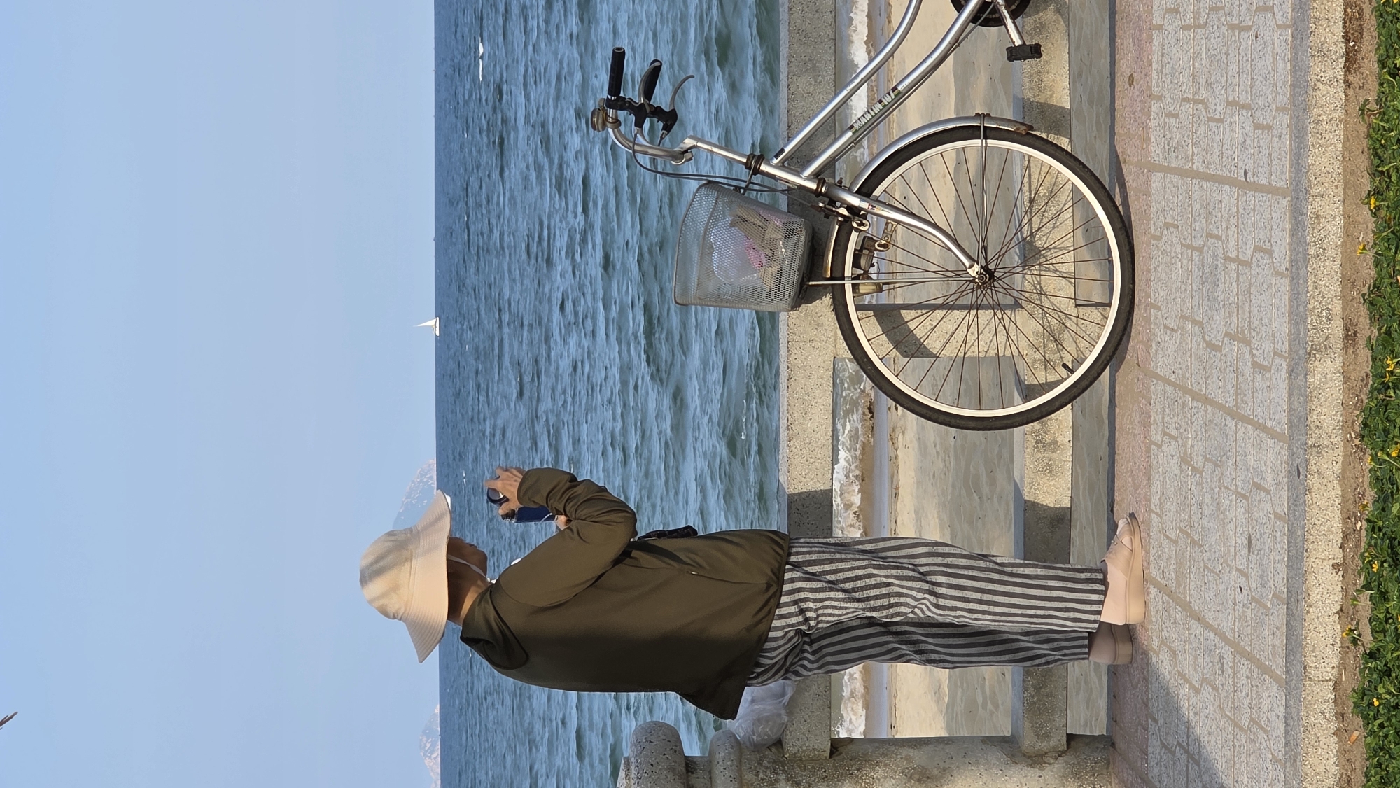 A person wearing an olive green jacket, striped pants, a wide-brimmed hat, and tan shoes is standing on a light-colored stone walkway taking a picture with a phone. A bicycle with a front basket is parked next to the person, partially blocking the view of the sea. The sea is visible in the background, with a sailboat on the horizon. The sky is clear and blue. The walkway has a low stone barrier, and there are steps leading down towards the sea. The location is likely in the city of Nha Trang, Vietnam.