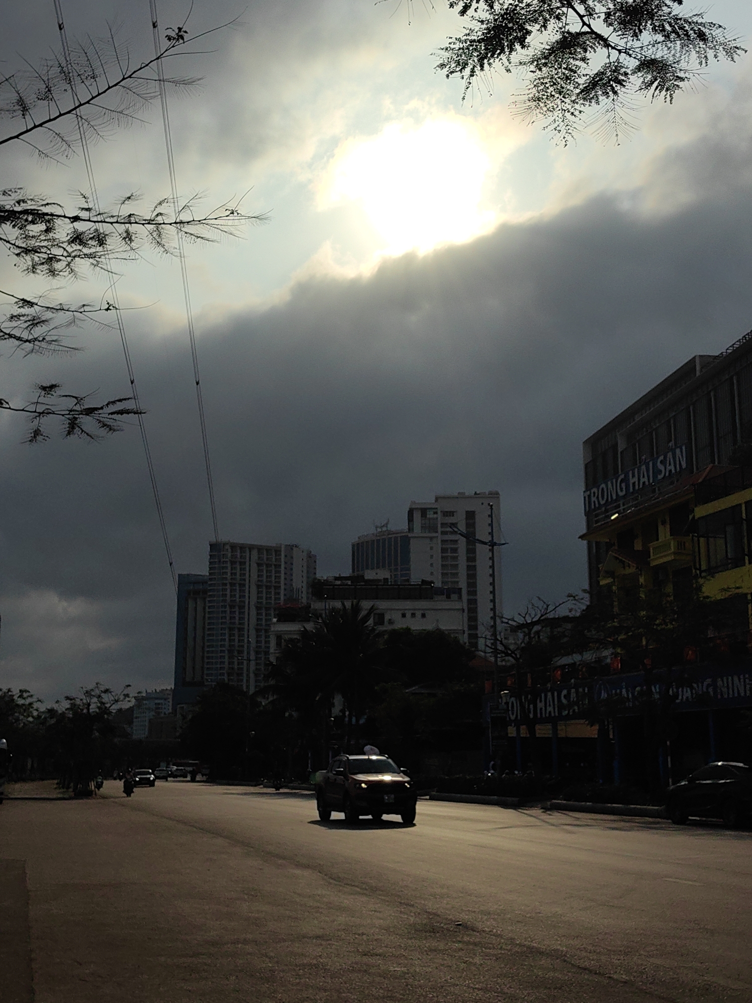 This vertical shot captures an urban street scene in Bai Chay Ward, Vietnam, under a dramatic sky. The image is characterized by strong backlighting, with the sun brightly breaking through heavy, dark grey clouds in the upper center, creating a high-contrast effect.
The foreground features a wide asphalt road, appearing somewhat rough or dusty, stretching into the distance. A dark-colored pickup truck with its headlights on (or reflecting the sun) is prominent on the right side of the road, driving towards the viewer. Other vehicles, including cars and motorbikes, are visible further down the road as dark silhouettes, indicating ongoing traffic.
To the right, a line of buildings flanks the street. One prominent building has a distinctive yellow lower section and features large white text on its facade: "TRONG HẢI SẢN". Below this, more text is partially visible, appearing to include "GIANG NINH". In the midground and background, several multi-story buildings, some appearing modern or under construction, rise against the dark sky, mostly obscured by deep shadows.
The upper left portion of the frame is dominated by silhouetted tree branches and slender utility lines, which crisscross against the lighter parts of the cloudy sky. Dark, lush vegetation lines the road on both sides, further emphasizing the dramatic lighting conditions. The overall mood is striking, with the intense natural light source creating a powerful focal point amidst the shadowed urban environment. The time of day is likely either early morning or late afternoon, or perhaps a moment when the sun briefly pierced through an otherwise overcast sky. No specific people are clearly discernible, but the moving vehicles suggest typical daily activity.