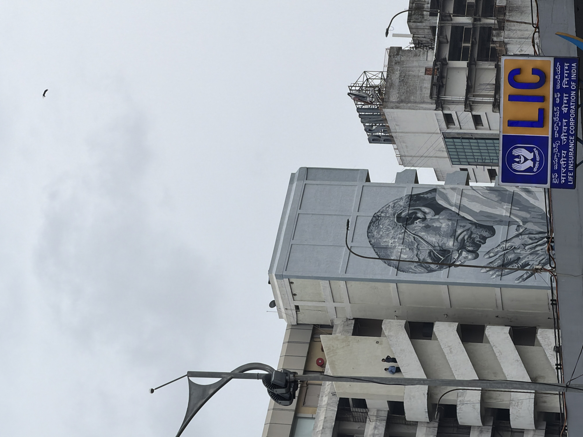 This media file shows a mural of Mahatma Gandhi on the side of a building in Hyderabad, India. The mural is black and white and depicts Gandhi's face and hands in a close-up view. Below the mural, there are several levels of balconies on the building, with two people visible on one of the balconies. In the foreground, there is a metallic structure with lights, possibly a street lamp or part of an overhead sign.
To the right of the building, a tall sign with "LIC" in blue letters on a yellow background is visible. Below the "LIC" logo, there is text in Telugu and Hindi, as well as English: "LIFE INSURANCE CORPORATION OF INDIA" and "भारतीय जीवन बीमा निगम" and "లైఫ్ ఇన్సూరెన్స్ కార్పొరేషన్ ఆఫ్ ఇండియా". The sky is overcast, suggesting a cloudy day. In the upper left corner of the image, a bird is flying against the sky. The overall scene captures a mix of urban architecture, art, and everyday life in Hyderabad.