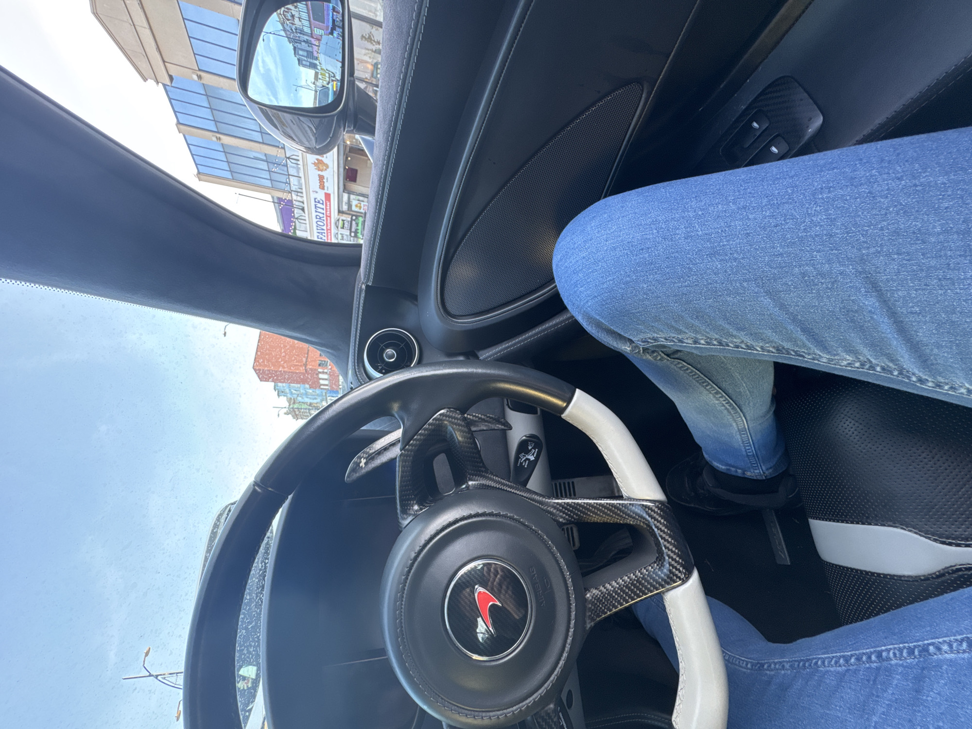 The image shows an interior view of a McLaren sports car with the driver seated, wearing blue jeans and black shoes. The steering wheel is black and white with carbon fiber accents and a McLaren emblem. Outside the car, buildings are visible, including a shop with the sign "Favorite" visible in the car's side mirror. The location appears to be Great Yarmouth, United Kingdom, based on the shop name and architectural style of the buildings in the background. The sky is visible through the windshield, appearing partly cloudy. The interior of the car is primarily black with leather and carbon fiber finishes.