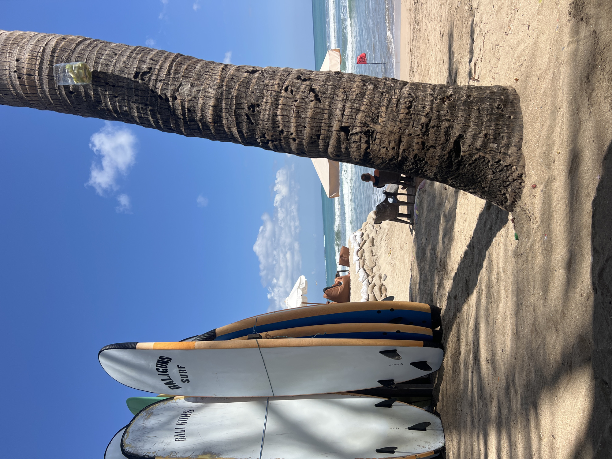 The image captures a sunny day on a beach in Kuta, Indonesia, likely near a surf school or rental shop. In the foreground, several surfboards are leaning against a rack. Two of the boards prominently display the text "BALIGUNS SURF". The boards are white with colorful accents and black fins. A large palm tree trunk occupies the right side of the frame, casting a shadow across the sandy beach.
In the middle ground, the ocean waves are gently rolling towards the shore under a clear blue sky with scattered white clouds. Two beach chairs are placed on the sand, facing the ocean. A person, seen from the back, is sitting in one of the chairs. Behind the chairs, a light-colored beach umbrella provides shade, and a pile of sandbags forms a low barrier. A small red flag, possibly indicating surf conditions or a designated swimming area, is planted on the beach near the water.
Further in the background, the coastline extends, and the ocean stretches to the horizon. The overall scene conveys a relaxed, tropical atmosphere.