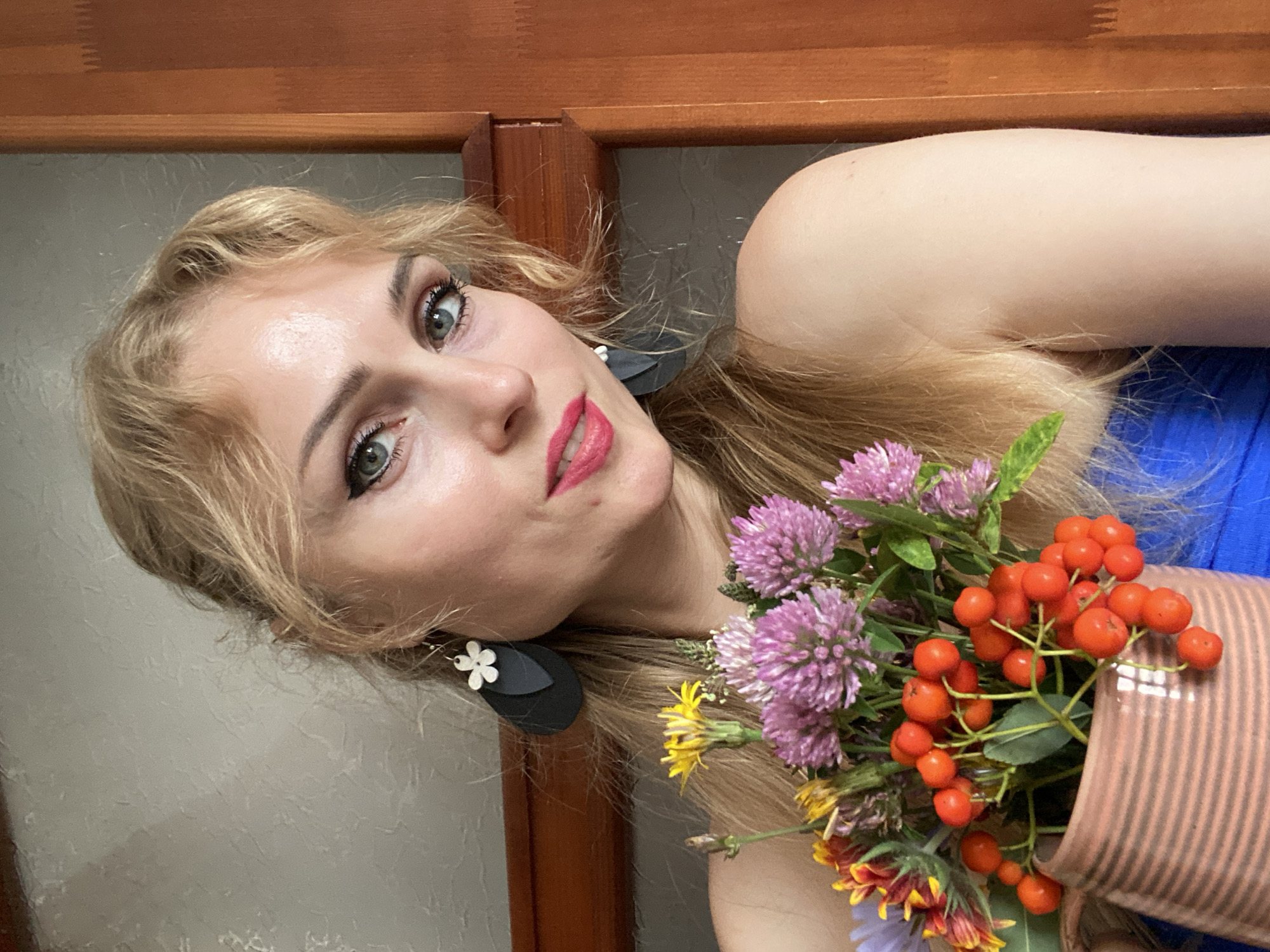 A blonde woman with green eyes and red lipstick holds a vase of flowers. She is wearing a blue top and black earrings. The background appears to be an indoor setting with a wall and a wooden door. The image was taken in Lida, Belarus.