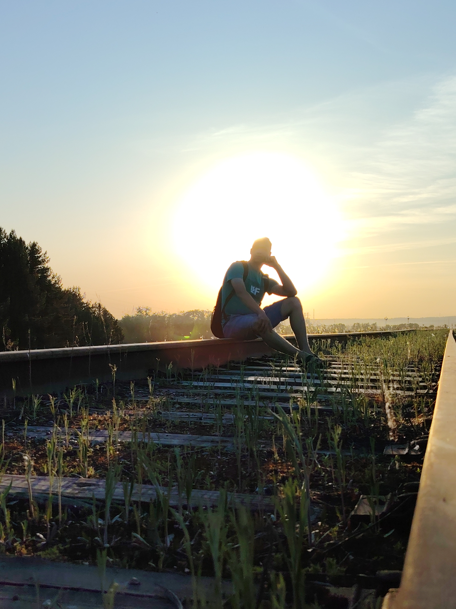A medium shot depicts a person sitting on a railroad track against a bright sunset, located in Chaikovsky, Russia. The person is seated with their legs dangling, and their right elbow is resting on their knee while their hand supports their head. They are wearing a t-shirt, shorts, and shoes. The railroad tracks are covered with vegetation. The bright sun creates a halo effect around the person, obscuring details. The background includes trees and industrial structures.