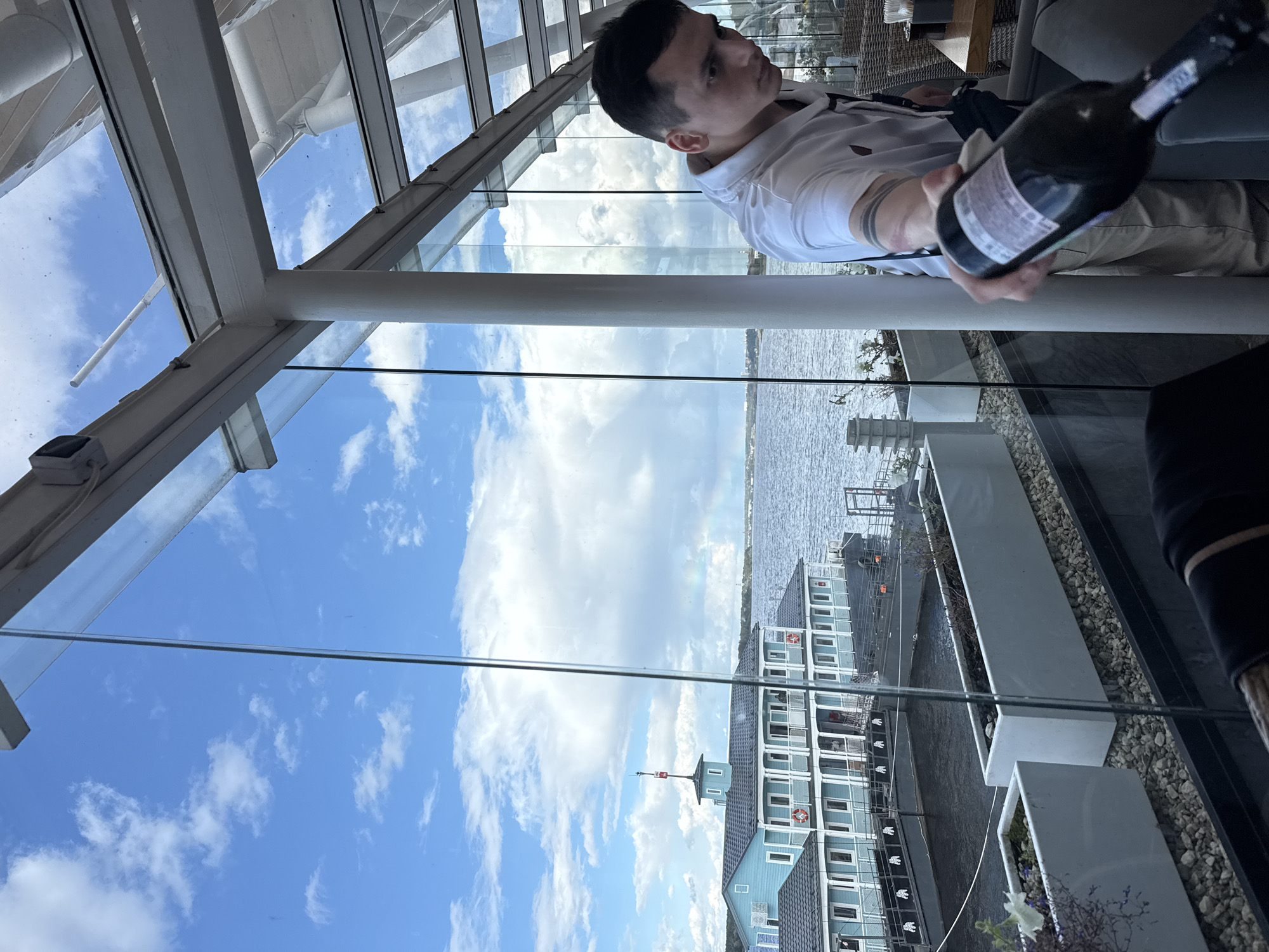 A man is holding a dark bottle of wine in a restaurant with a view of a body of water and a light blue building with white trim. The man is wearing a white collared shirt and light-colored pants. He has a tattoo on his left arm. The restaurant appears to be enclosed by glass walls and a glass roof. In the background, there is a building on the water with multiple windows and a flag pole. The sky is blue with white clouds. The scene suggests a meal or gathering at a waterside establishment. This establishment is located in the city of Perm, Russia, on the Kama River embankment, likely at the location of the former port and the "Stolichnaya" restaurant.