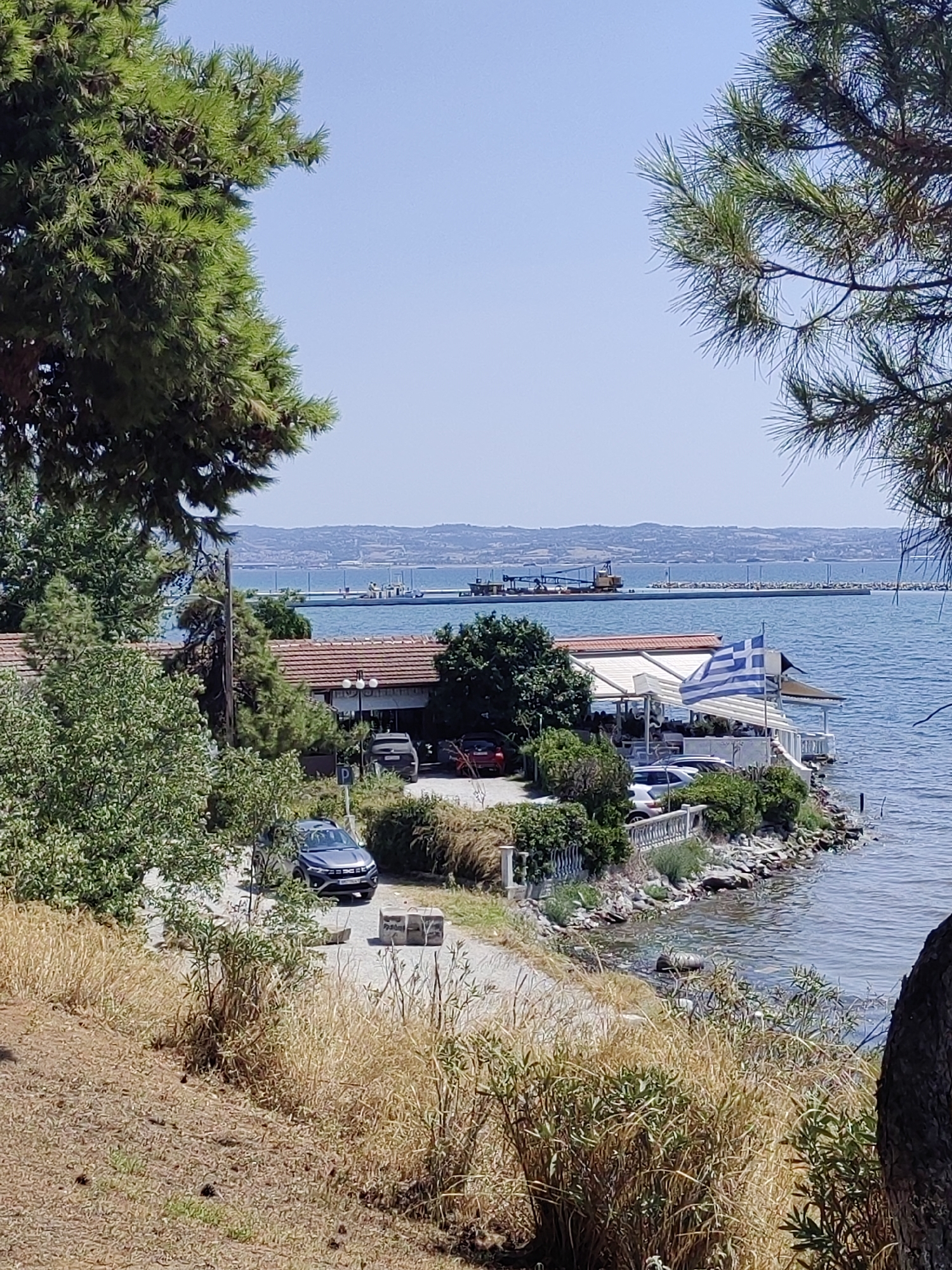The image depicts a waterfront scene, likely a seaside restaurant or cafe, with a Greek flag prominently displayed. The location is on the coast of Thessaloniki, Greece, offering a view of the Aegean Sea. In the foreground, dry grasses and trees frame the scene, with a pathway leading to a parking area where several cars are visible, including a dark gray SUV.
The restaurant itself features a light-colored awning and outdoor seating areas. Beyond the restaurant, the sea stretches out to a distant coastline, with a long pier or breakwater equipped with industrial structures, possibly a crane, suggesting port activity. The water is calm, reflecting the clear blue sky. The overall impression is of a sunny day at a coastal establishment.