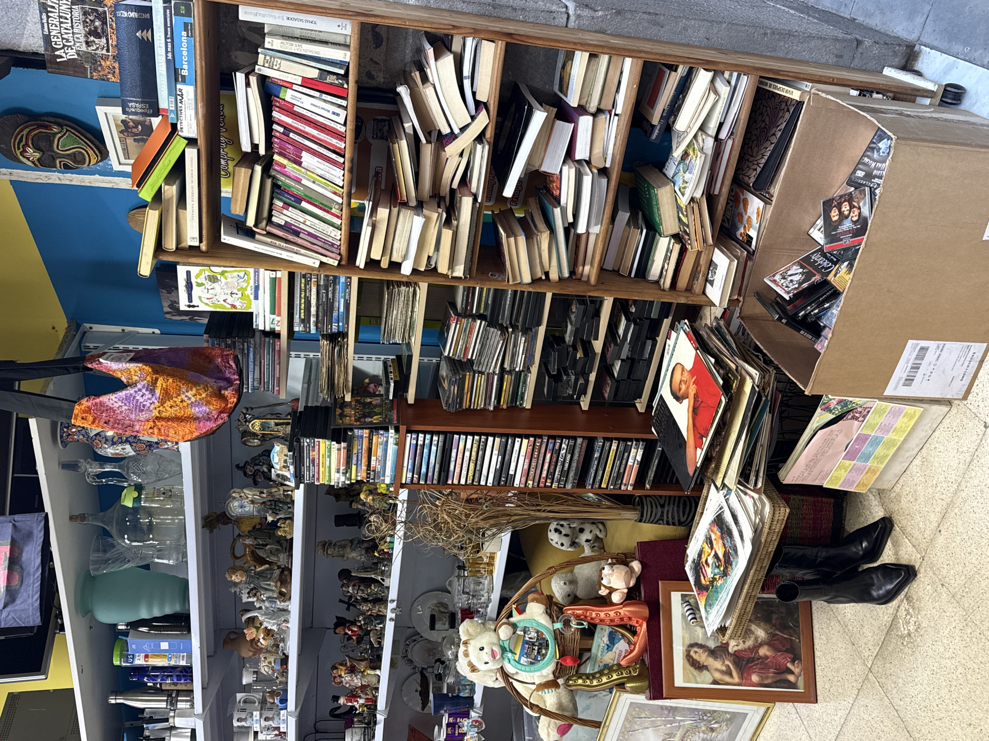 The image depicts an indoor second-hand shop likely located in Barcelona, Spain, given a book titled "Barcelona" visible in the image. There's a crowded bookshelf filled with books and DVDs. A cardboard box overflowing with DVDs is precariously stacked next to the bookshelf. Below the bookshelf, there is another shelf with glassware, figurines, and other decorative items. Boots are on the floor among the items. The scene conveys an atmosphere of clutter and a diverse collection of pre-owned items.