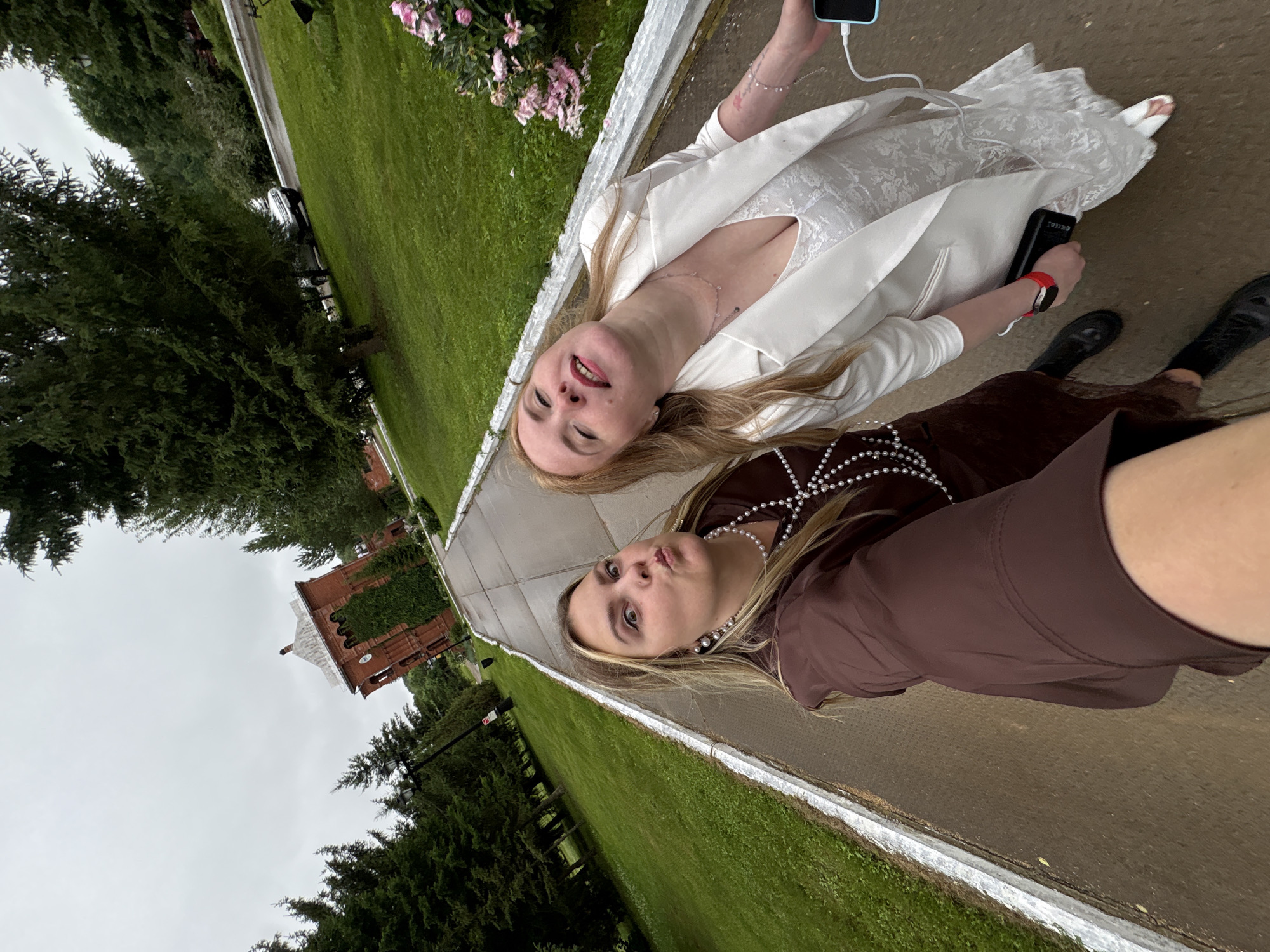 The image shows two young women posing for a selfie outdoors in Zavod Mikhailovskii, Russia. One woman on the left wears a white suit jacket over a lace dress and holds a light blue smartphone. She has blonde hair and is smiling with her eyes closed, appearing happy. The other woman on the right wears a brown dress with a pearl necklace and holds a black phone with a red smartwatch on her wrist. She has blonde hair and is making a playful "fish face" expression.
The setting is a paved walkway bordered by a green lawn and trees. In the background, a red brick building with ivy growing on its walls is visible, and a car is parked on the side of a road. The sky is overcast, suggesting it might be a cloudy day. The overall mood is casual and lighthearted, as the women are enjoying themselves and capturing a moment together.