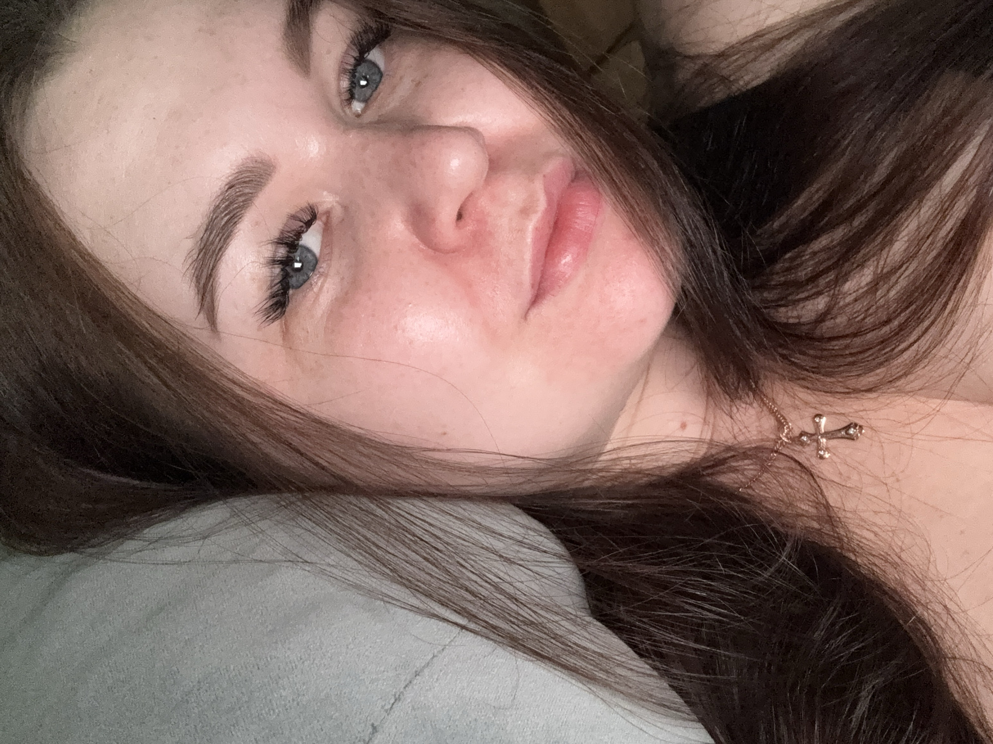 The image is a close-up, selfie-style photograph of a young woman with long, brown hair. She has striking blue eyes, prominent eyelashes, and her eyebrows are well-defined. Her skin appears fair with some redness, possibly from blushing or skin condition, and a few freckles are visible. She is wearing a delicate gold necklace with a cross pendant. The background is out of focus, but appears to be a soft, light-colored surface, possibly a pillow or blanket. The lighting is soft and warm, casting a gentle glow on her face. The image does not provide any information about the location in the city of Chaikovsky, Russia.