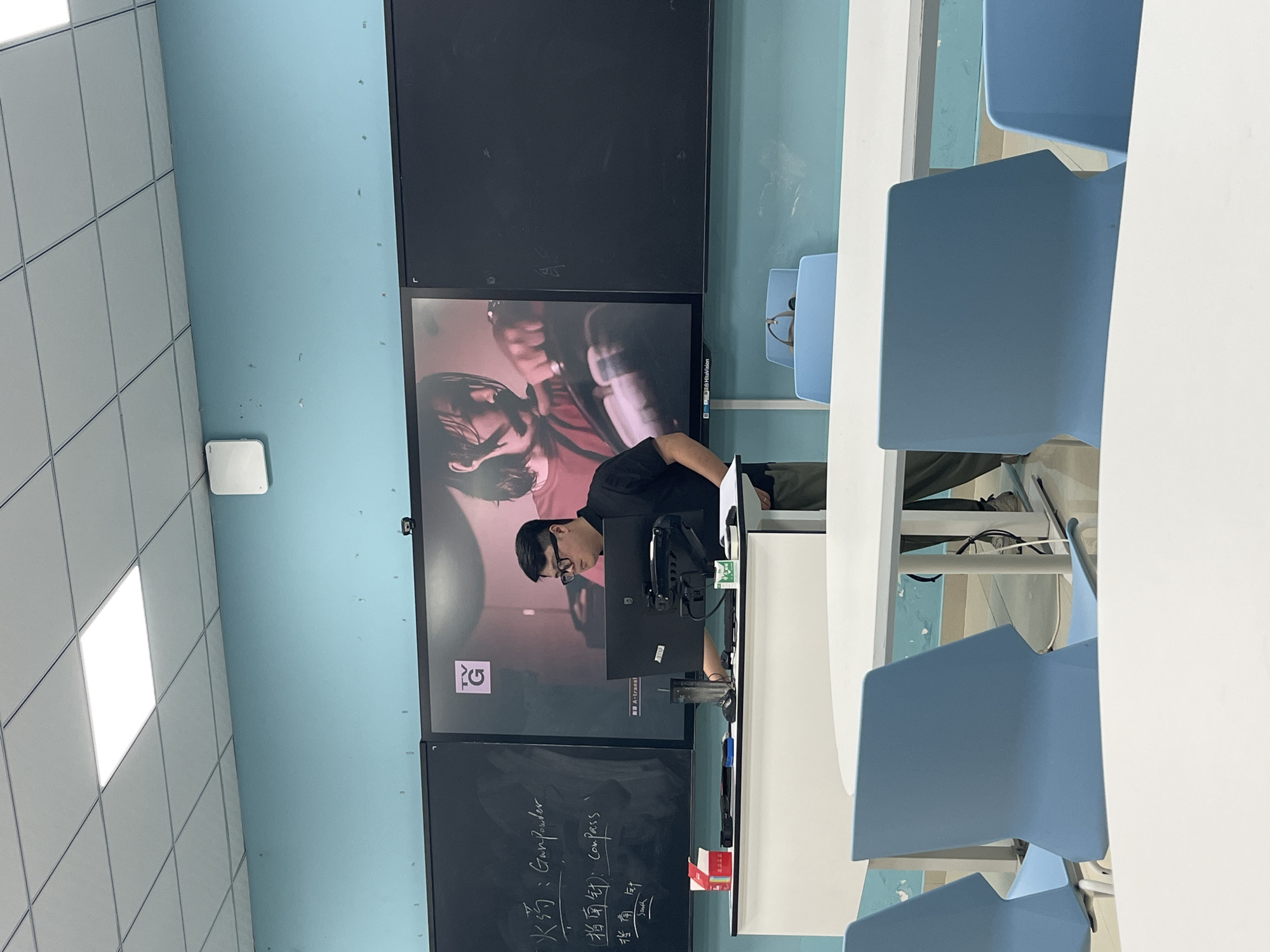 The image depicts a classroom setting where a man wearing glasses and a black shirt is standing behind a podium, interacting with a computer. A large screen behind him displays an image of a person with dark hair wearing a red shirt. To the left of the screen, a blackboard contains handwritten Chinese characters and their English translations, including "火药: Gunpowder," "指南针: Compass," and "指南针 South." The classroom features light blue walls, a white tiled ceiling with fluorescent lighting, and several white oval tables and light blue chairs arranged for students. This scene is identifiable as being located within the city of Jiangjin, China, likely within an educational institution.