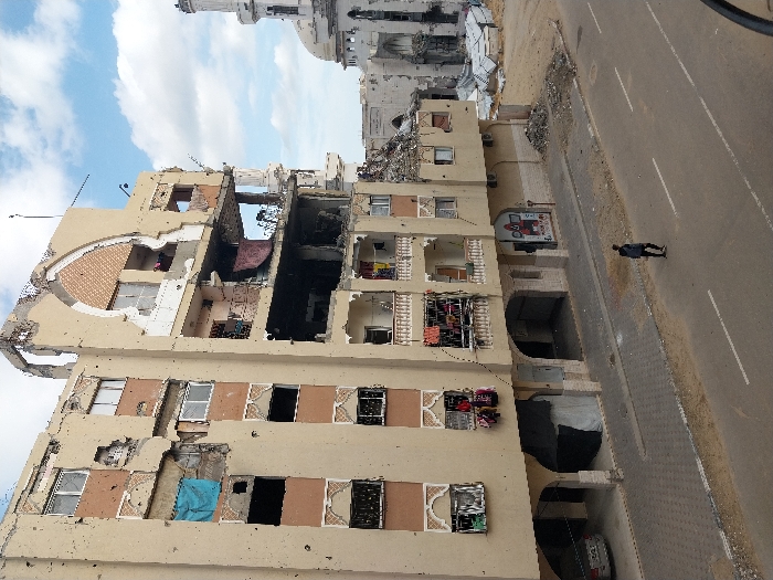 The image depicts a street scene in Khan Yunis, Palestinian Territory, characterized by significant structural damage to buildings under a partially cloudy sky. In the foreground, a paved asphalt road with white markings runs horizontally, where a lone male figure in dark clothing walks away from the viewer towards the background. To the left, a multi-story building, at least five floors high, exhibits extensive destruction. Its light beige facade is pockmarked with numerous impact points, and large sections of walls and windows are missing, revealing interior spaces and exposed rebar, particularly on the upper floors where an arched decorative element is partially destroyed. Clothes hang from some of the remaining balconies and out of shattered window frames. The ground floor features arched entryways, with a parked silver car under one and a dark tarp covering another. Adjacent to this building's right, in the background, stands a severely damaged mosque, identifiable by its partially intact dome and a ruined minaret. Debris and rubble are scattered on the unpaved ground surrounding the mosque.